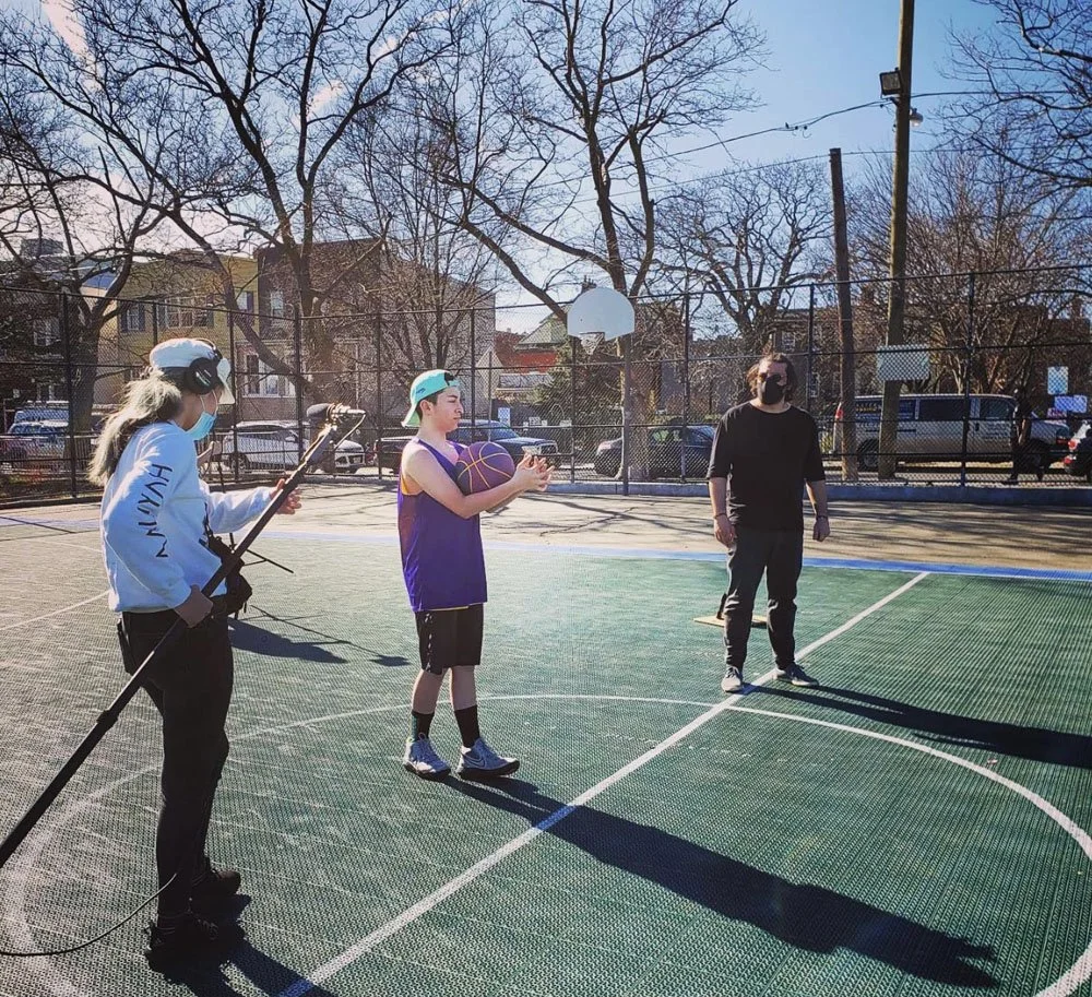 A woman and a girl are on an outdoor basketball court with a man standing nearby. The girl is holding a basketball, and the woman is recording with a camera on a tripod. The scene is sunny with leafless trees and parked cars in the background.