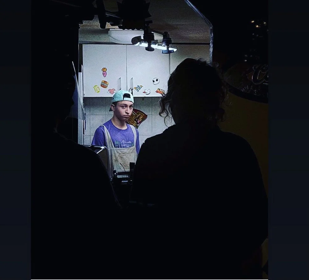 A boy standing in a kitchen with a serious expression, facing forward, wearing a cap backwards and an apron, seen through a silhouette of two people in the foreground.