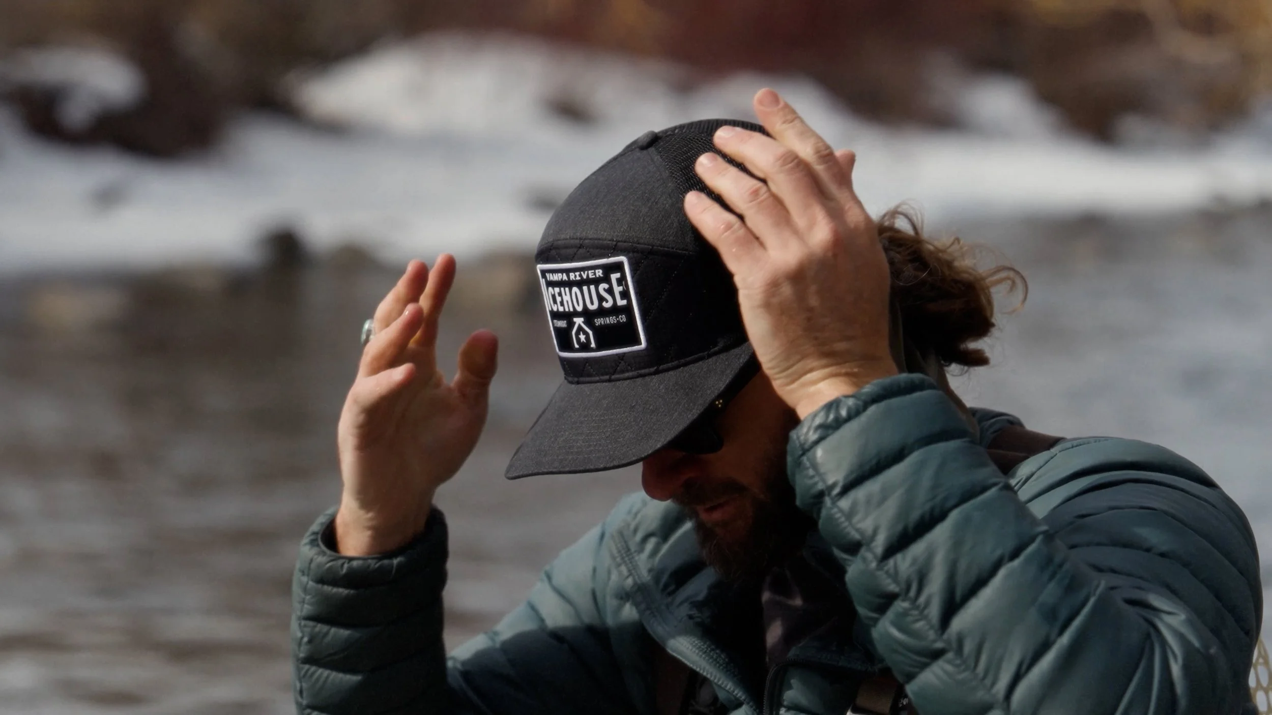 A person with long hair and a beard adjusting a black cap that says 'Vampire River Icehouse' while standing outdoors near a body of water.