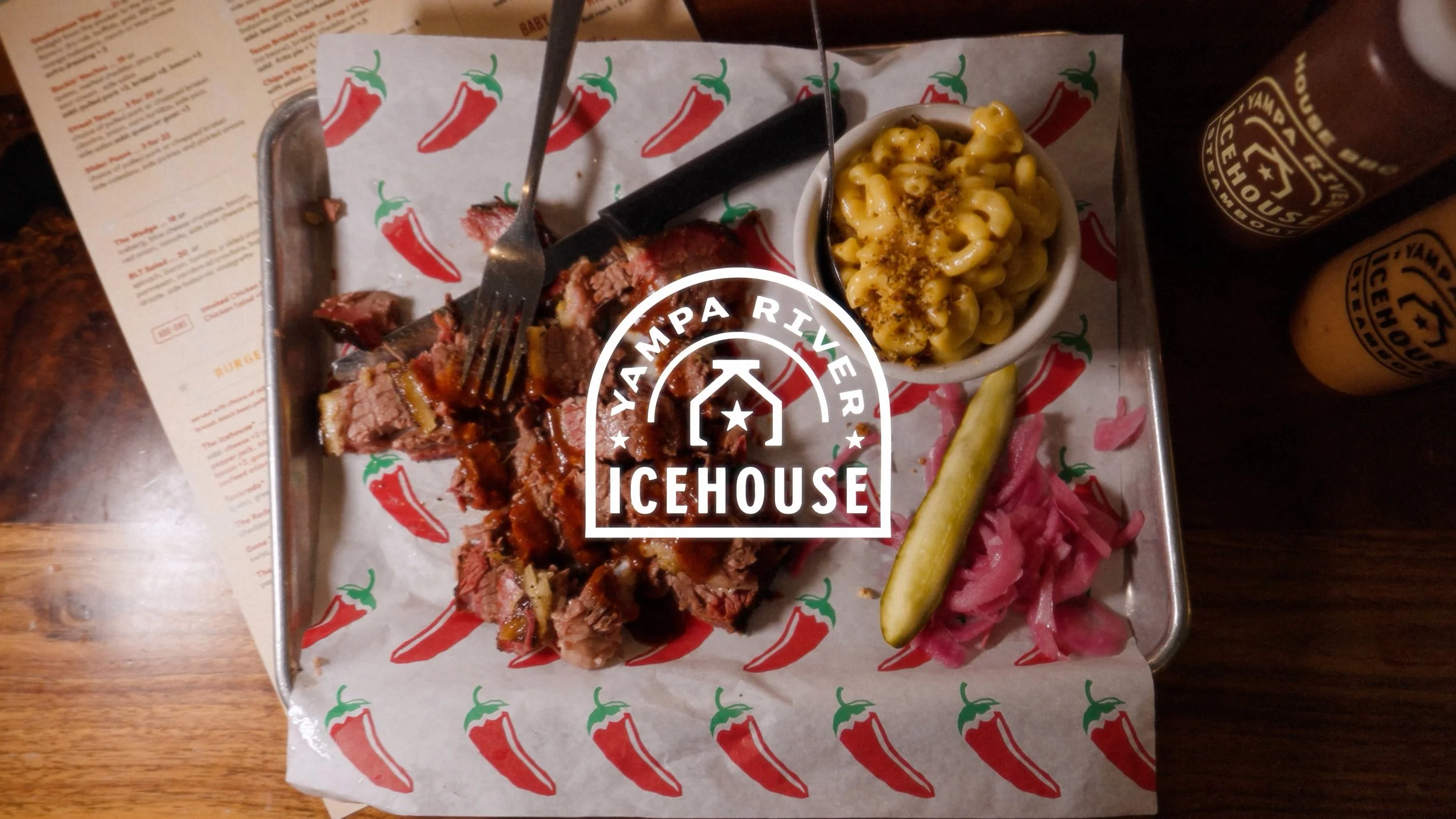 A tray of food from a Yampa River Icehouse restaurant includes sliced beef with sauce, a pickle spear, pink pickled onions, macaroni and cheese, and hot sauce bottles, with a paper liner decorated with red chili peppers.