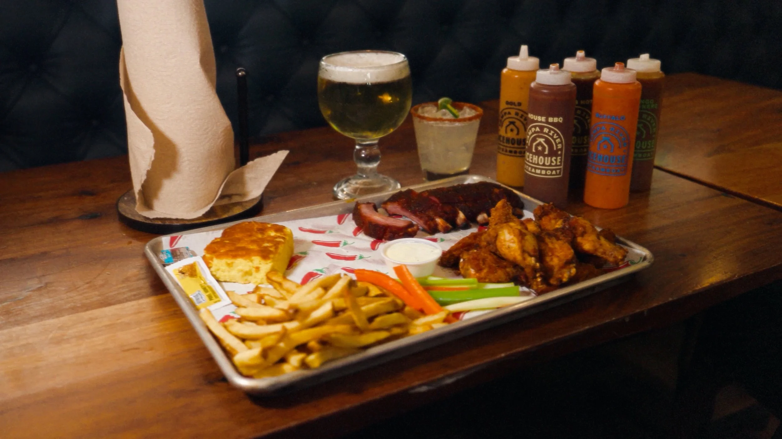 A tray of barbecue food including chicken wings, sliced brisket, macaroni and cheese, French fries, and vegetable sticks with dipping sauce. There is a glass of beer, shot glass, and four bottles of hot sauce on a wooden table.