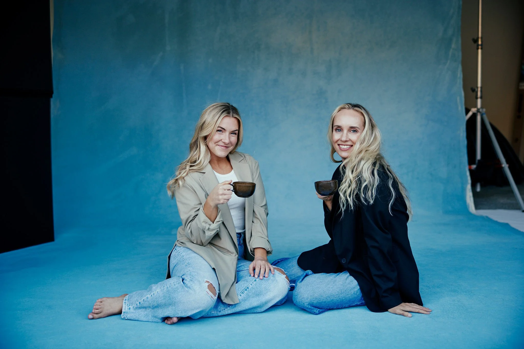 Two women sitting on the floor in front of a blue backdrop, holding coffee cups and smiling at the camera.