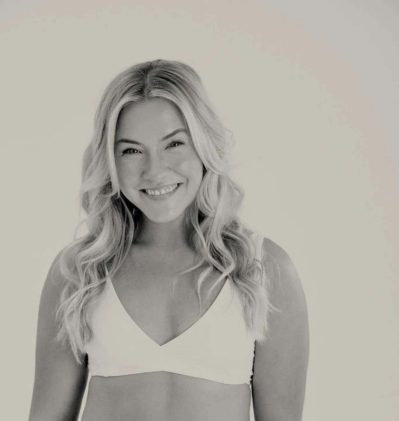 Black and white photo of a smiling woman with long, wavy hair wearing a white top, standing against a plain background.