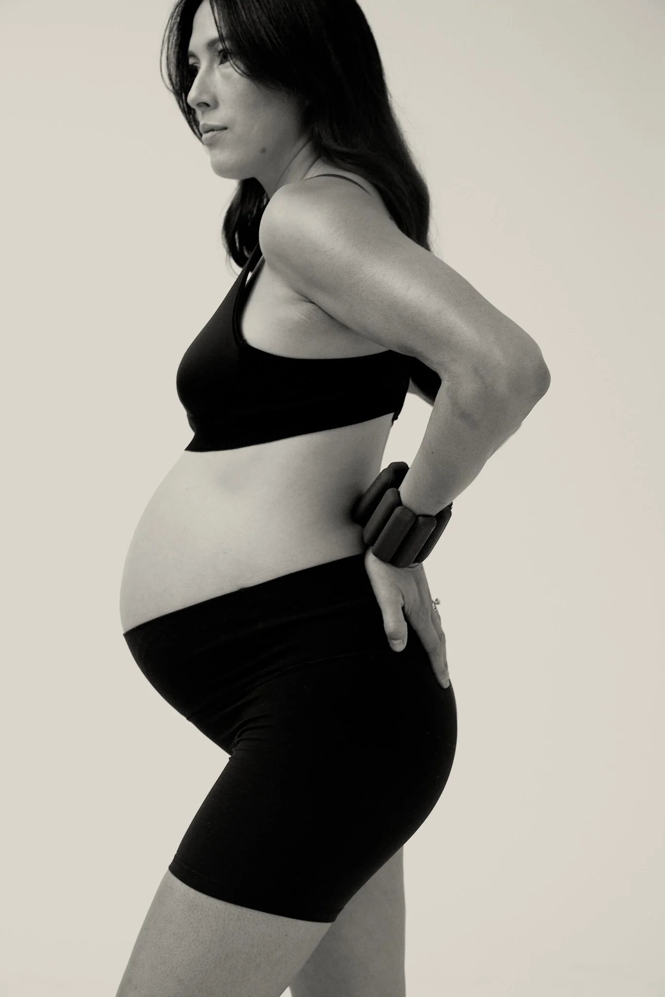 Black and white photo of a pregnant woman in athletic wear, standing sideways with one hand on her back and the other resting on her hip, looking to the side.