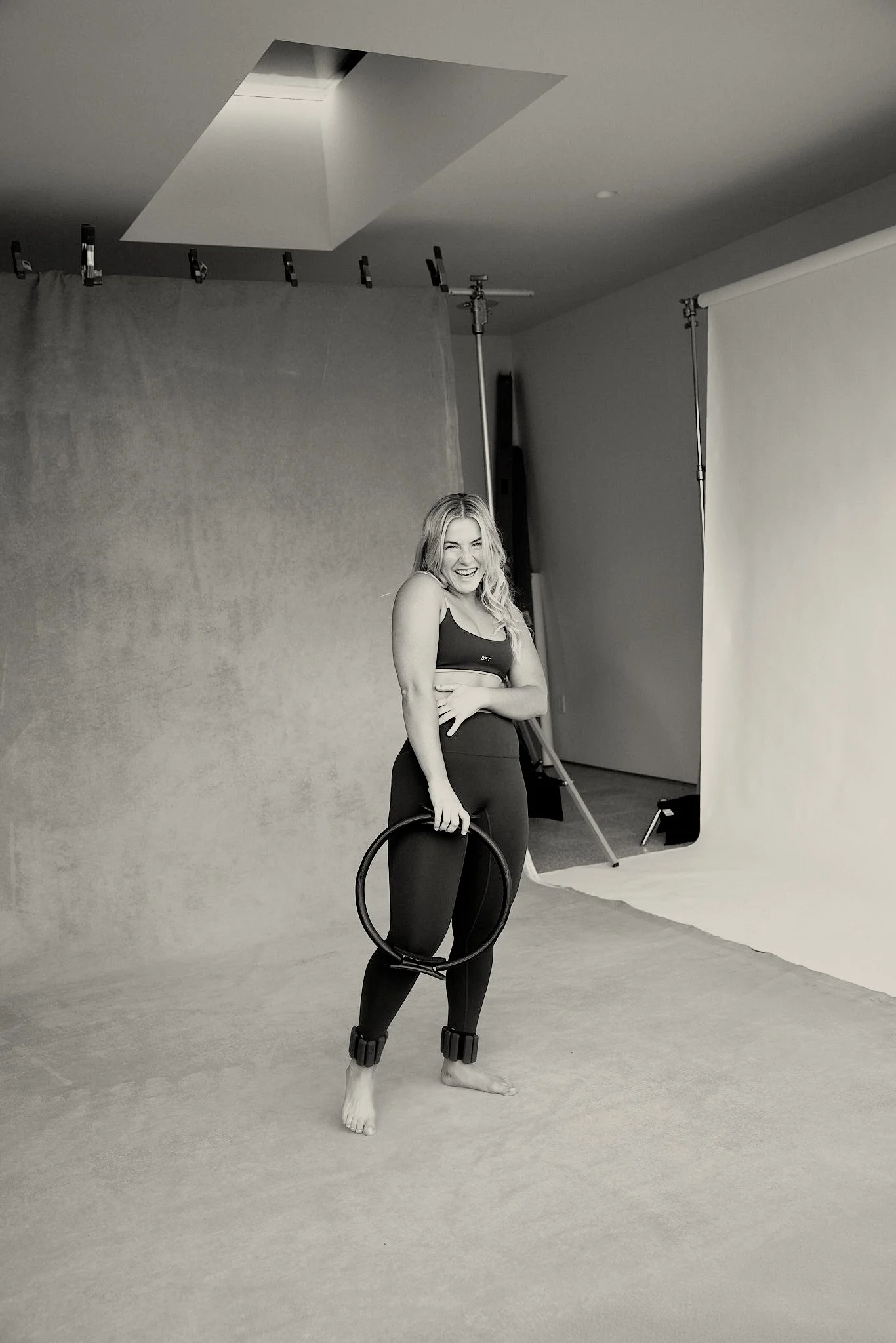 A woman standing on a photography studio set, holding a Pilates ring and laughing. She is dressed in workout clothing with ankle weights, and the set includes a backdrop and studio equipment.