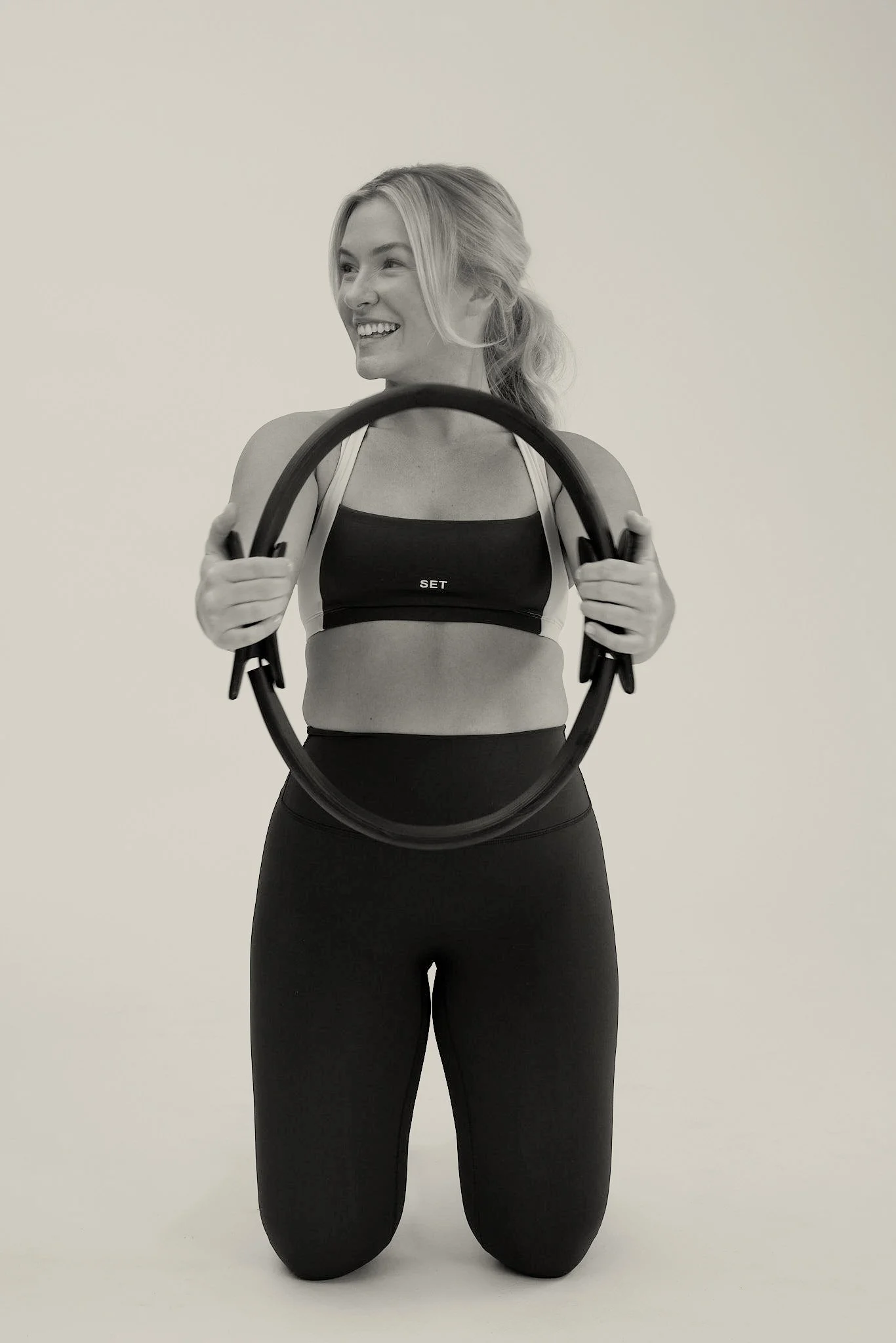A woman kneeling on the floor holding a Pilates ring, smiling and looking to her left, against a plain background, in workout attire.