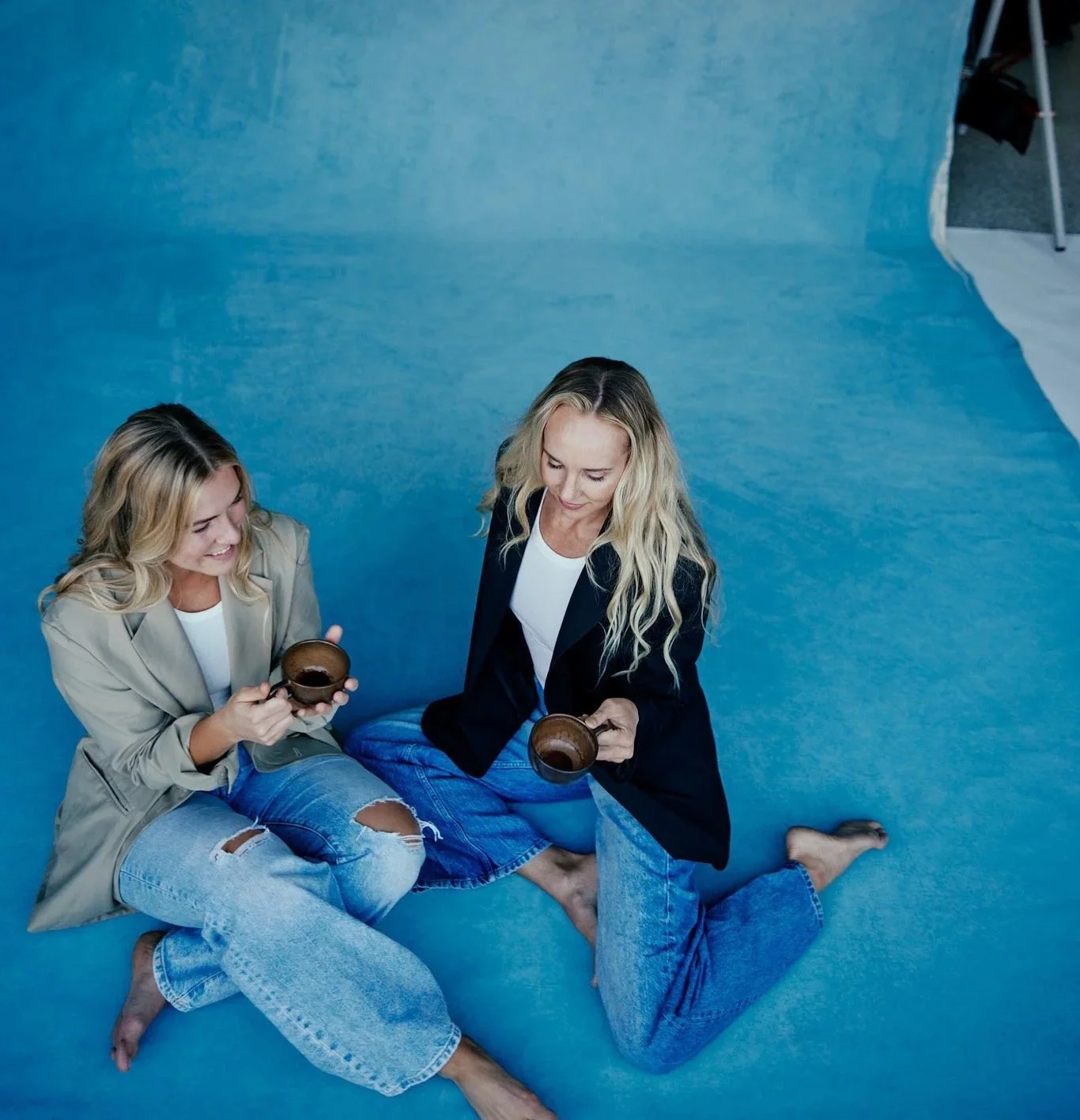 Two women sitting cross-legged on a blue floor, smiling and holding cups of coffee.
