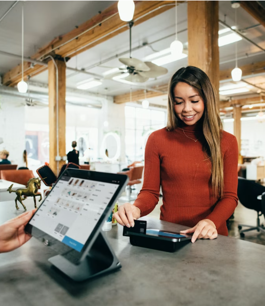 Una mujer sonriendo en una cafetería o espacio de trabajo, usando una tarjeta de pago o dispositivo para realizar una compra frente a un asistente que usa una tableta para gestionar transacciones.