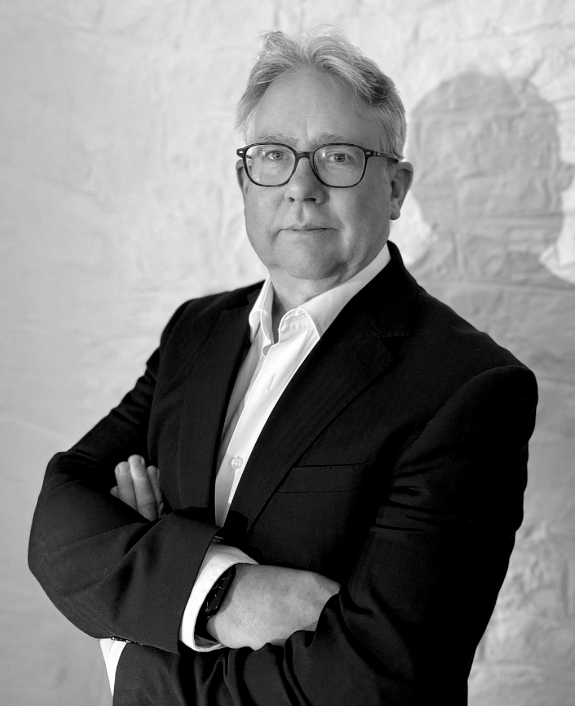 Black and white portrait of a middle-aged man with glasses, dressed in a suit jacket and white shirt, standing with arms crossed against a textured wall background.