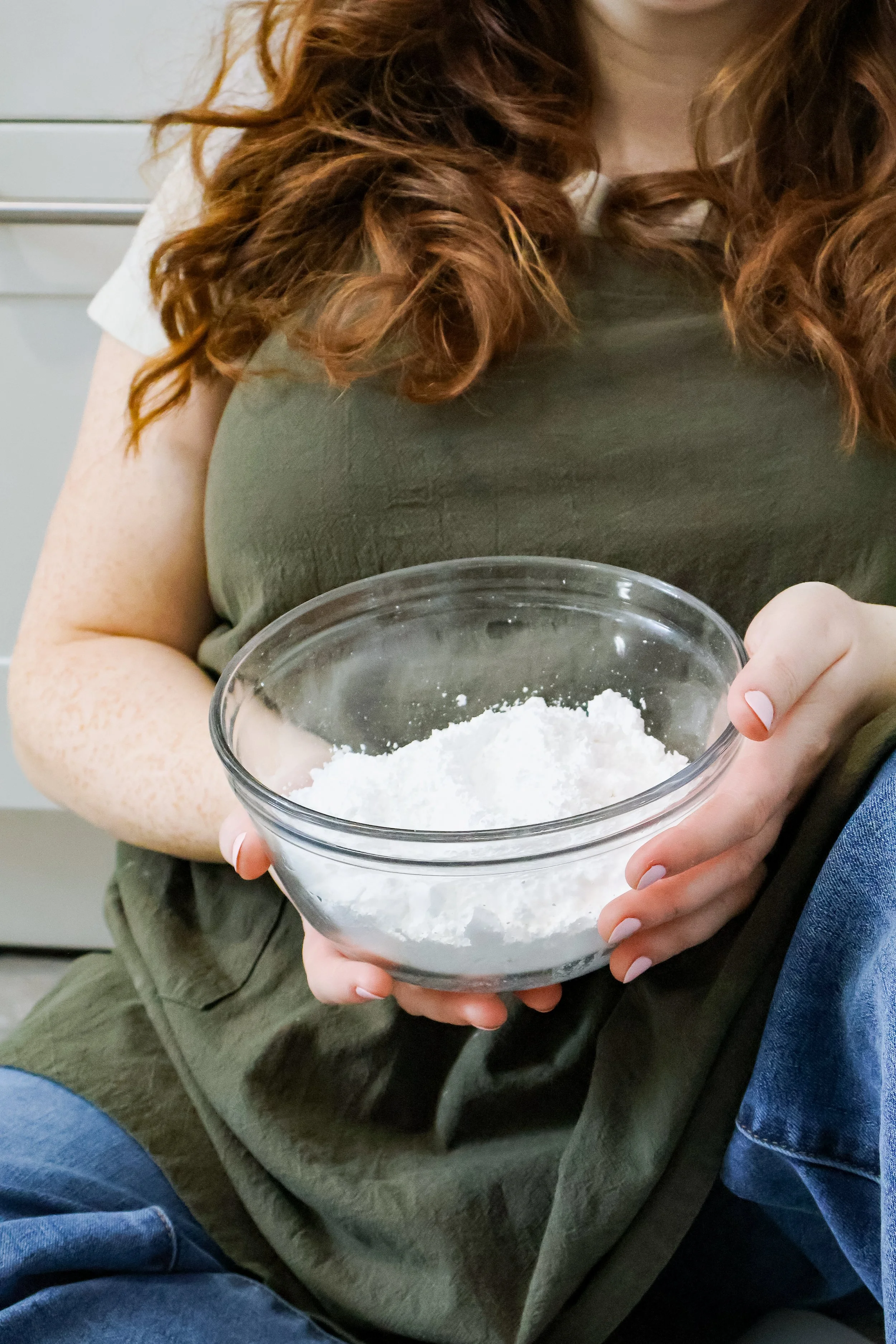 A woman with long red hair holding a glass bowl of white flour, sitting on a chair.