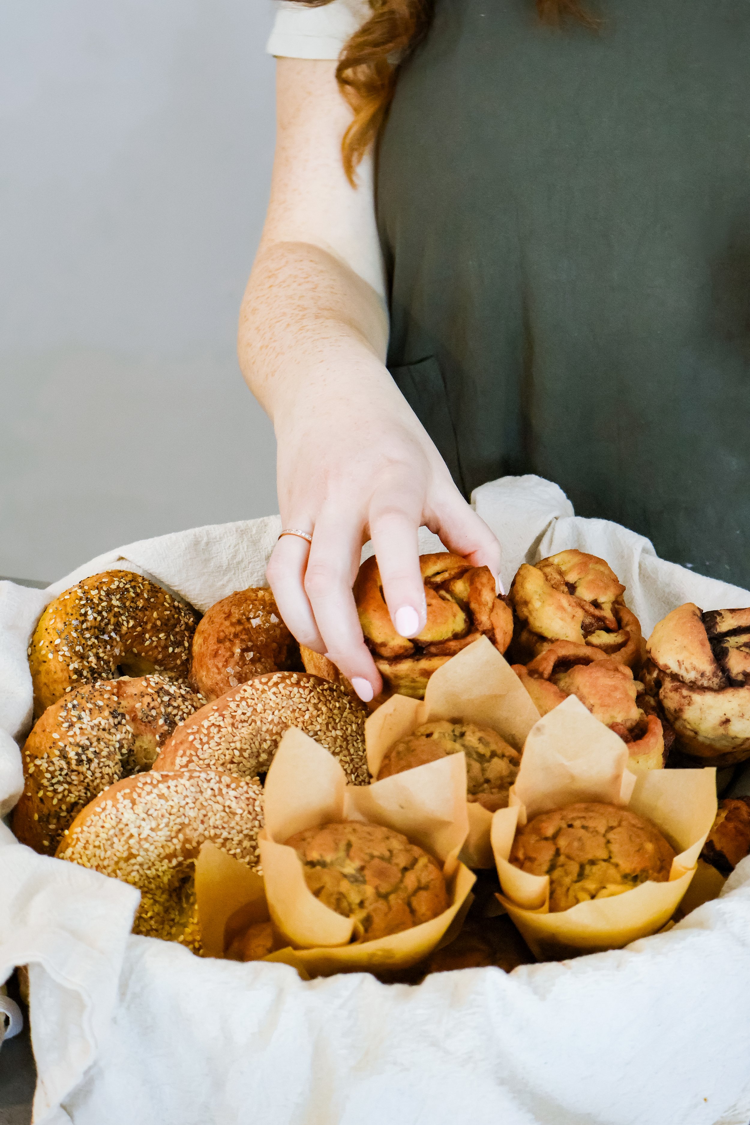 A person reaching into a basket filled with assorted baked goods, including muffins and bagels.