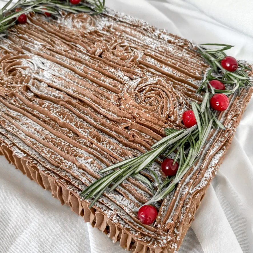 A large rectangular chocolate cake decorated with chocolate frosting, garnished with fresh rosemary sprigs and red cherries, dusted with powdered sugar.