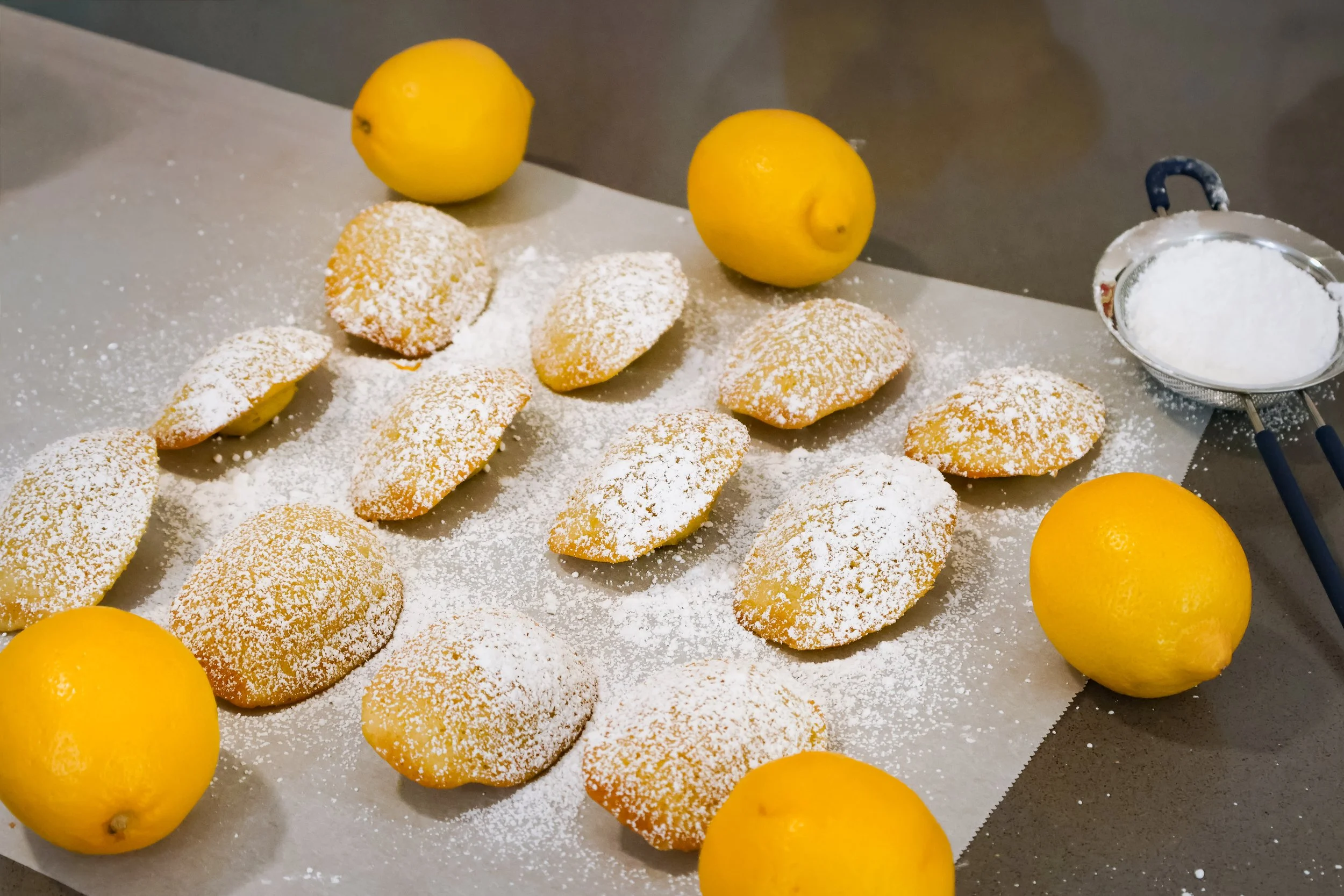 Lemon cookies dusted with powdered sugar on a baking sheet, surrounded by fresh lemons and a sifter of powdered sugar.