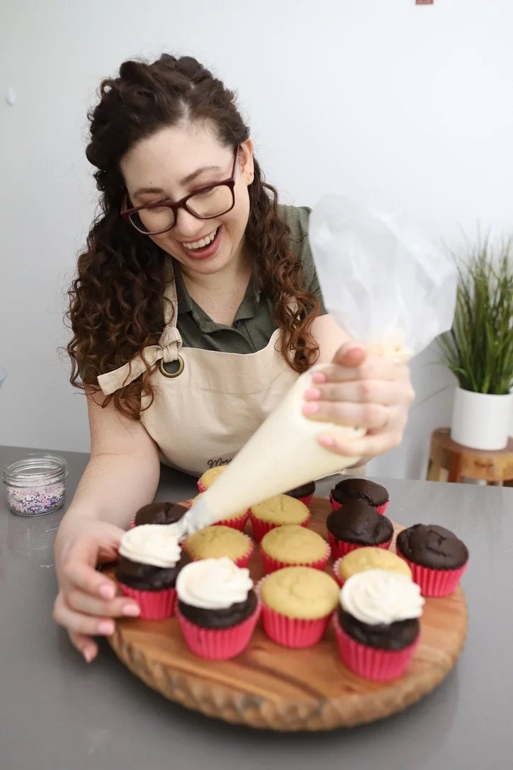 Woman in glasses and apron piping white frosting onto cupcakes on a wooden tray.