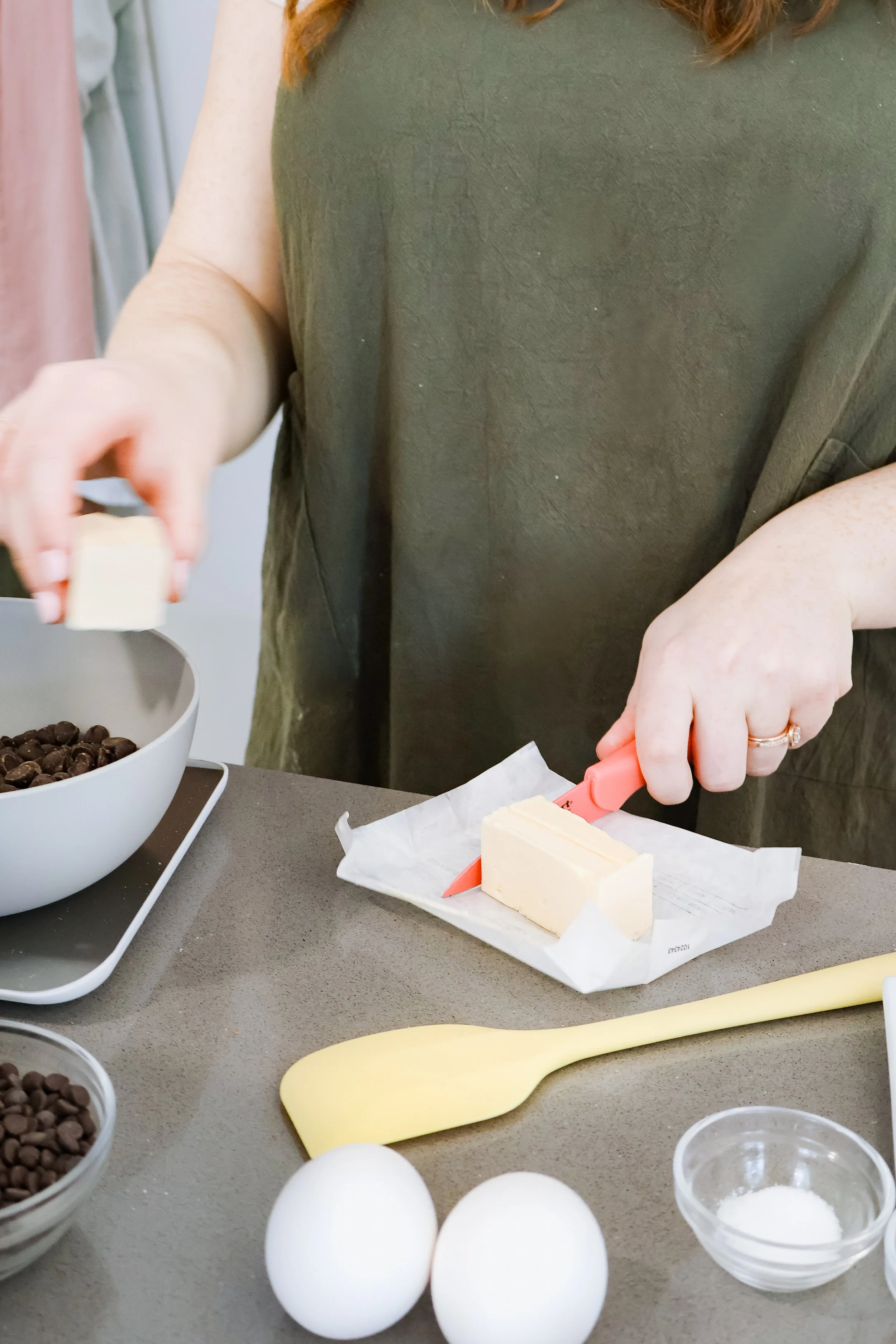 A person is slicing a stick of butter on a paper-lined surface in a kitchen, with eggs, chocolate chips, and small bowls of sugar nearby.