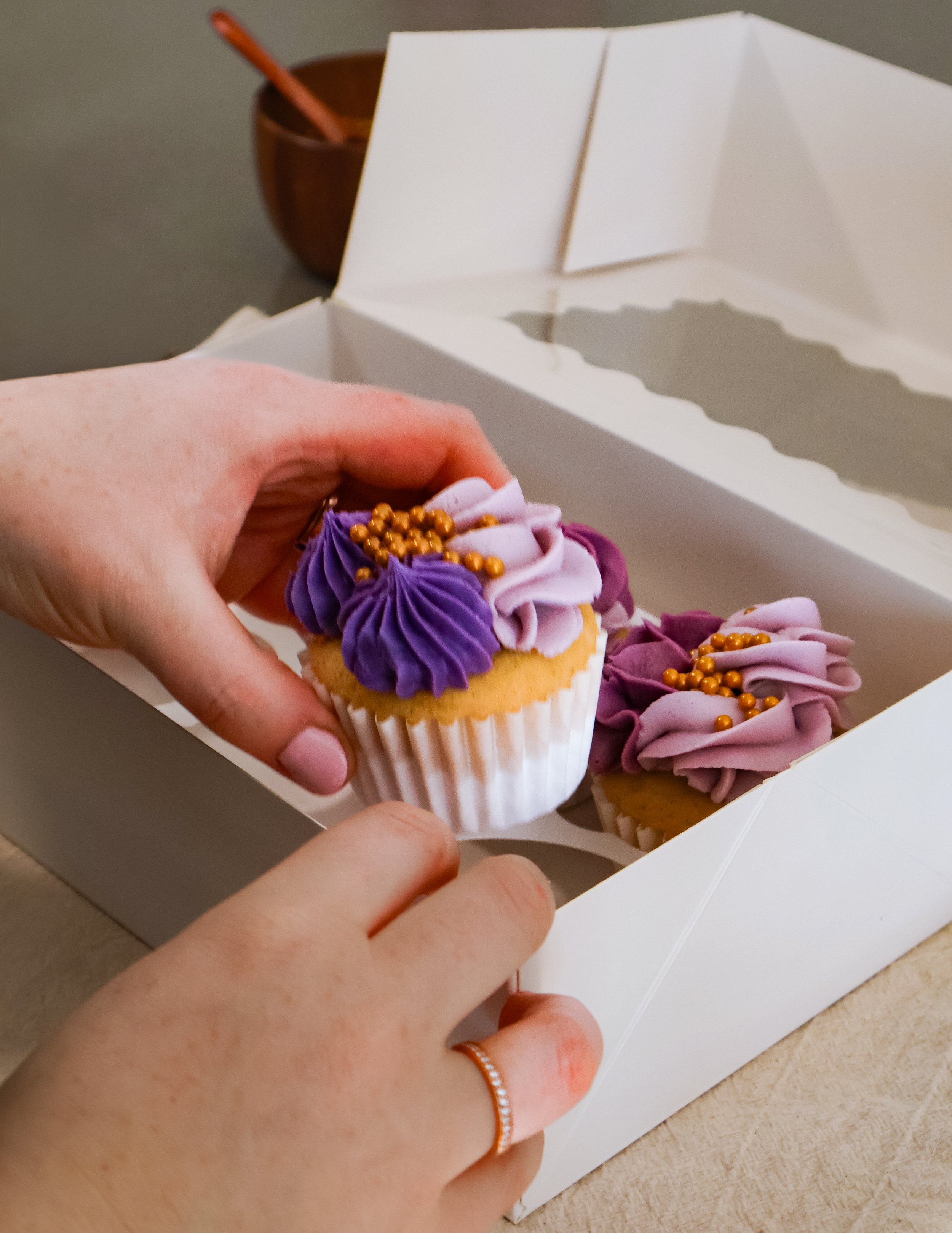 Person holding a cupcake with purple and pink frosting and gold beads, inside a white bakery box with two more cupcakes.
