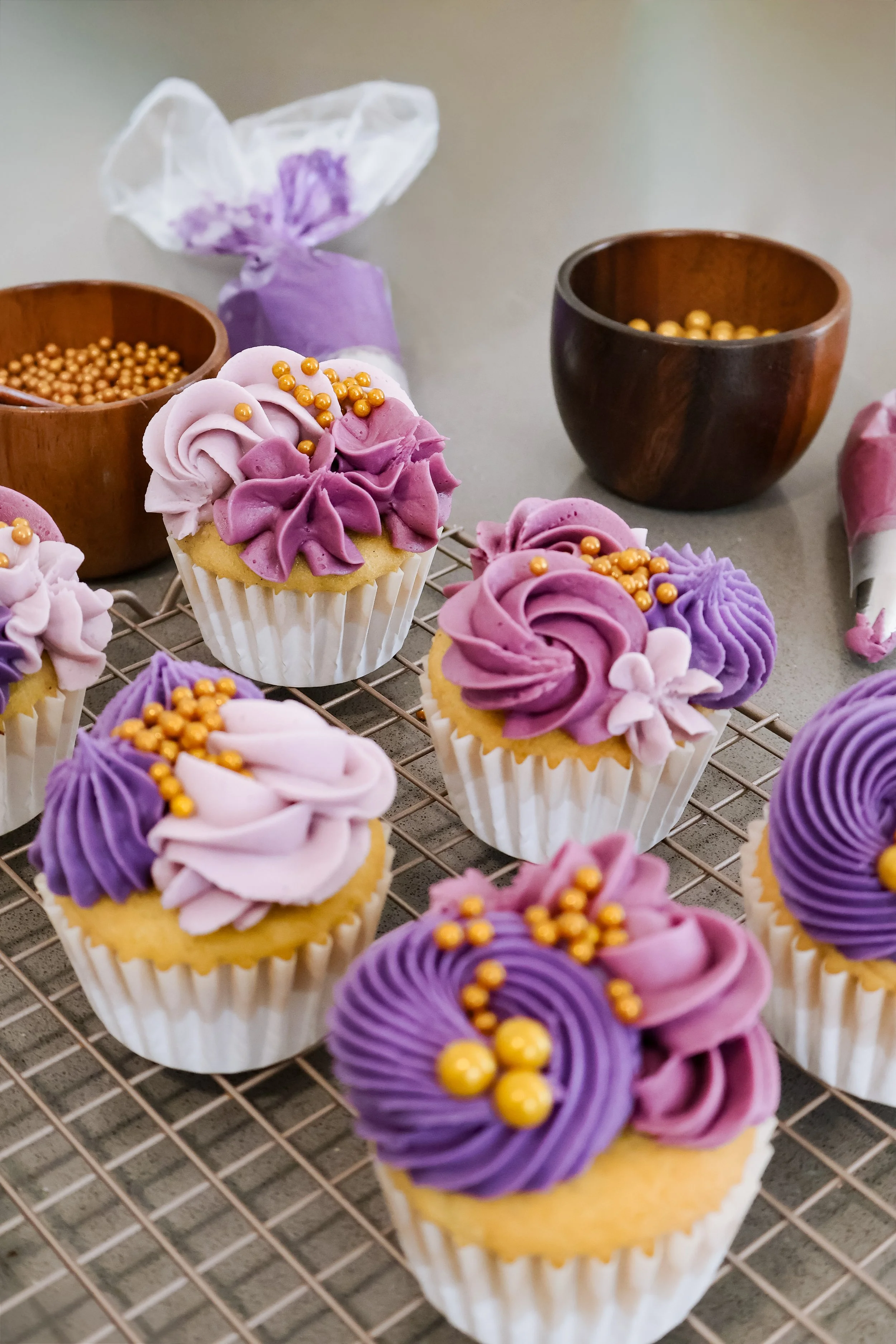 Purple decorated cupcakes with gold pearl and piped icing on a cooling rack, with icing and pearl items in the background.