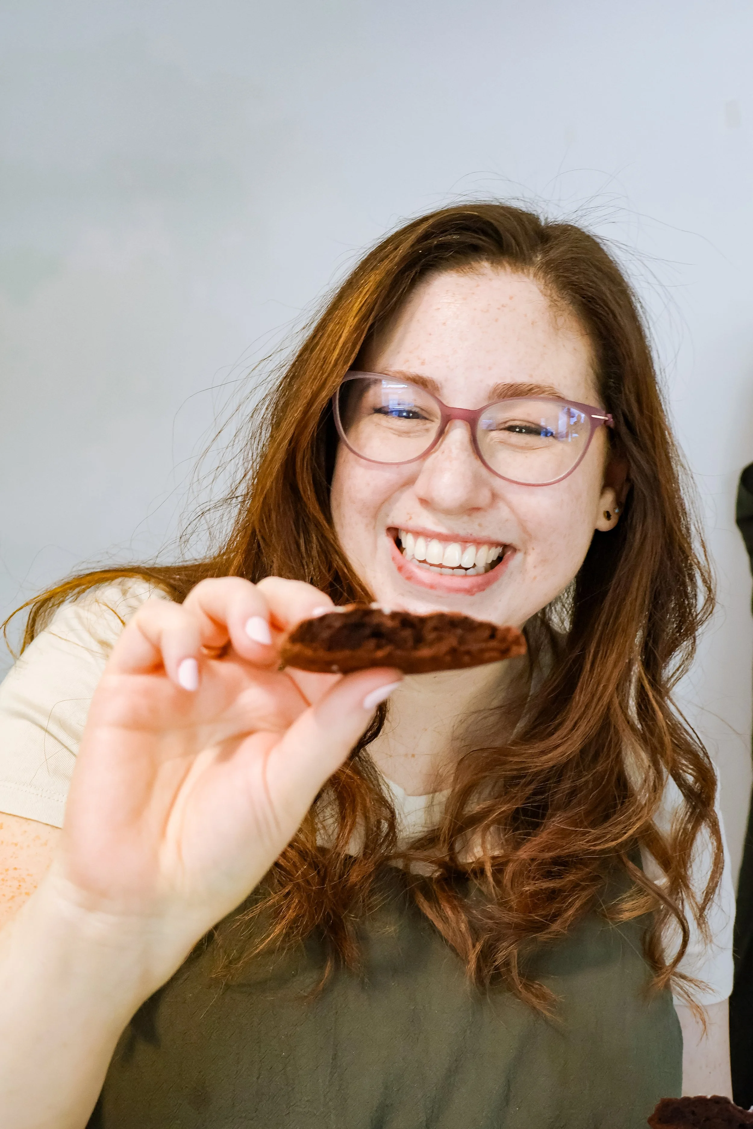 A smiling young woman with glasses and freckles holding a cookie towards the camera.