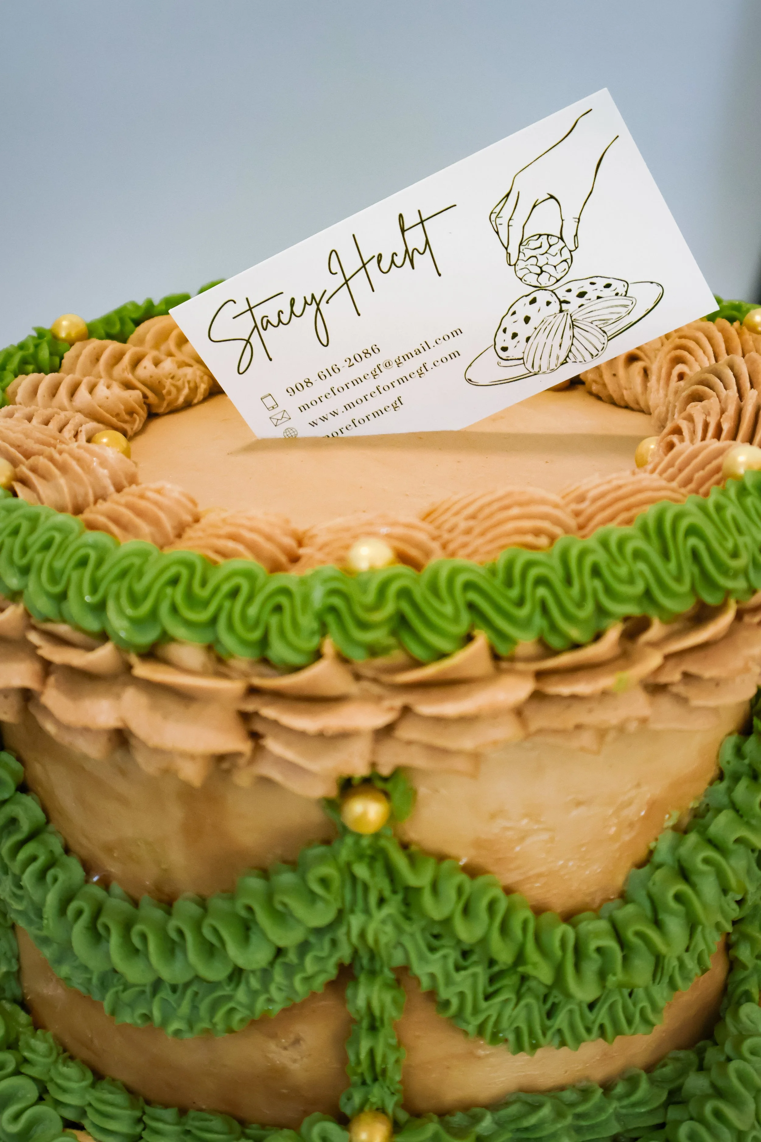 Close-up of a decorated cake with green and beige icing, and a business card on top that has a drawing of hand holding a coin and a plate of cookies, with contact information and name 'Stacy Hecht'.