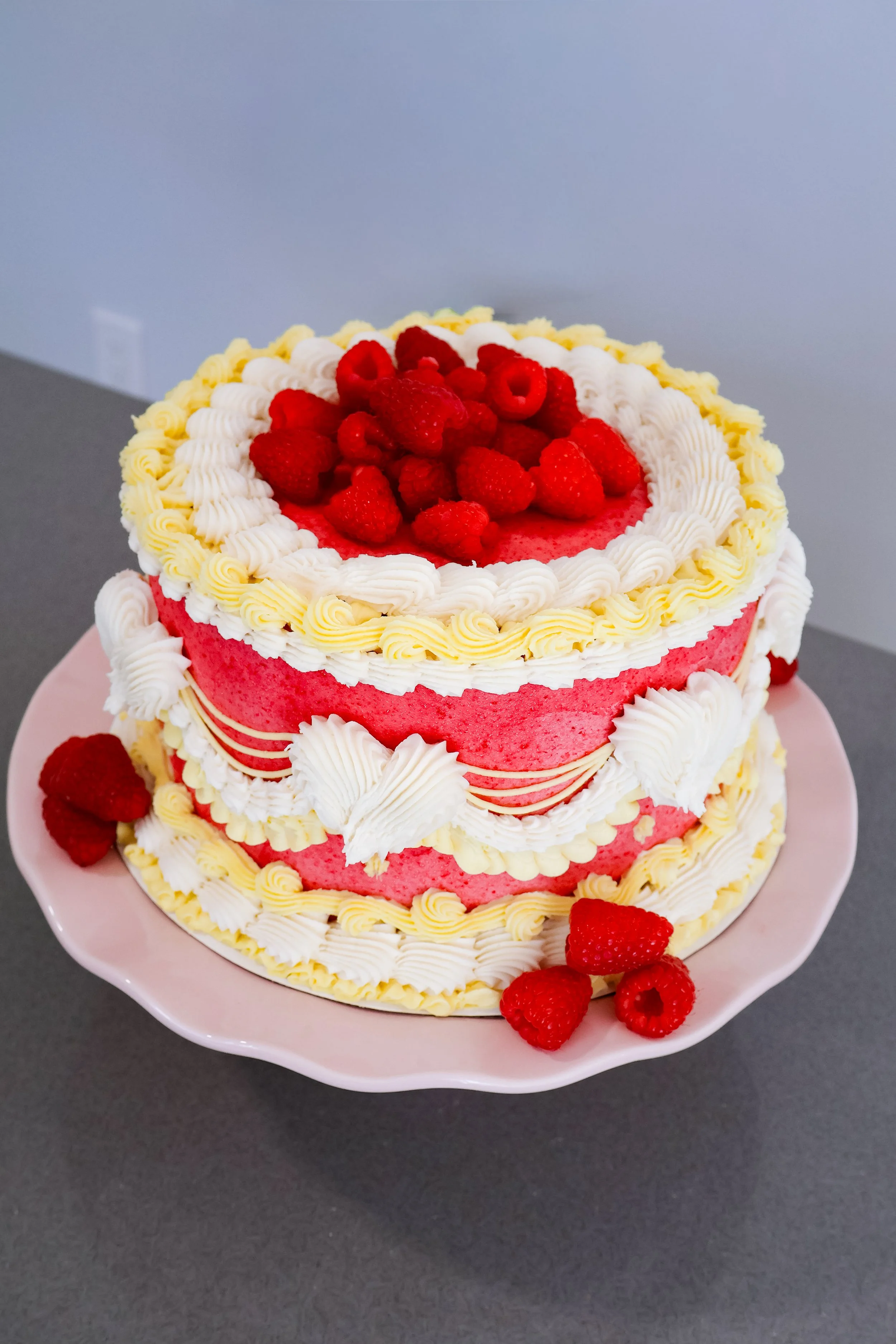 Round-shaped cake with pink frosting and yellow and white piping, decorated with raspberries