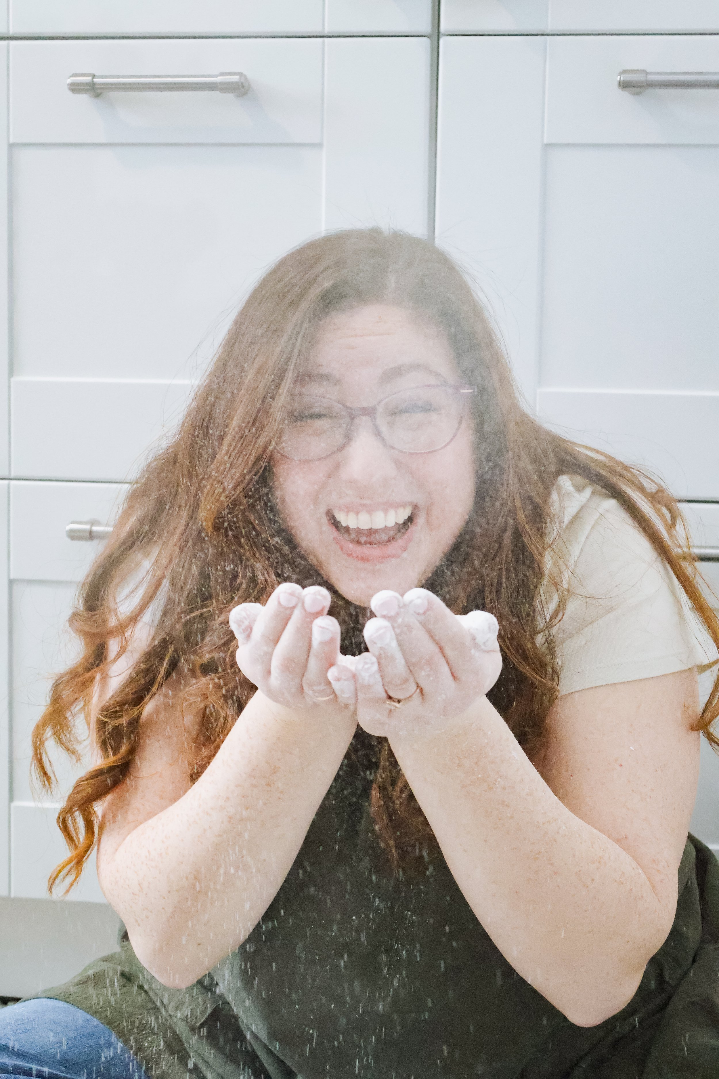 Woman in glasses and apron piping white frosting onto cupcakes on a wooden tray.