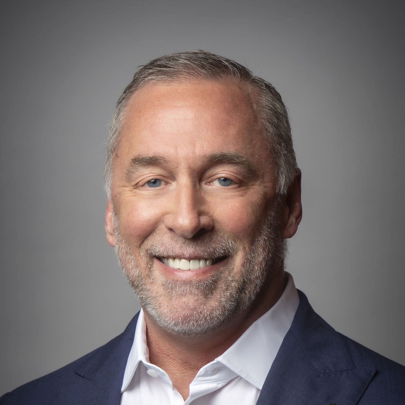 Headshot of a middle-aged man with gray hair and a beard, smiling, wearing a navy blazer and white shirt against a gray background.