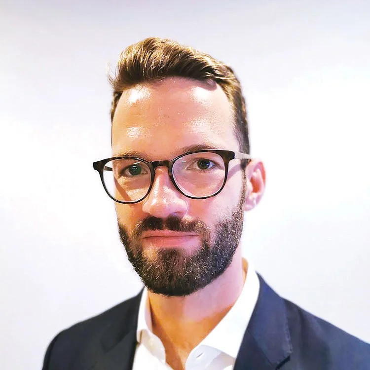 A man with glasses, a beard, and short hair in business attire, standing against a plain white background.
