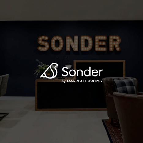 Interior view of a hotel lobby or reception area with a dark wall, illuminated letter marquee spelling "SONDER," and a desk with decorative flowers and clerical items.