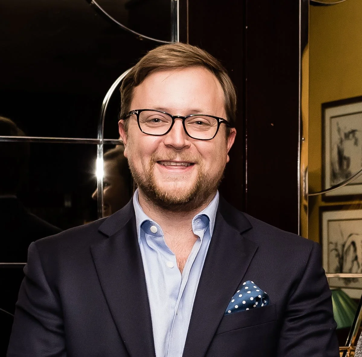 A smiling man wearing glasses, a navy blazer, and a light blue shirt, standing in an indoor setting.