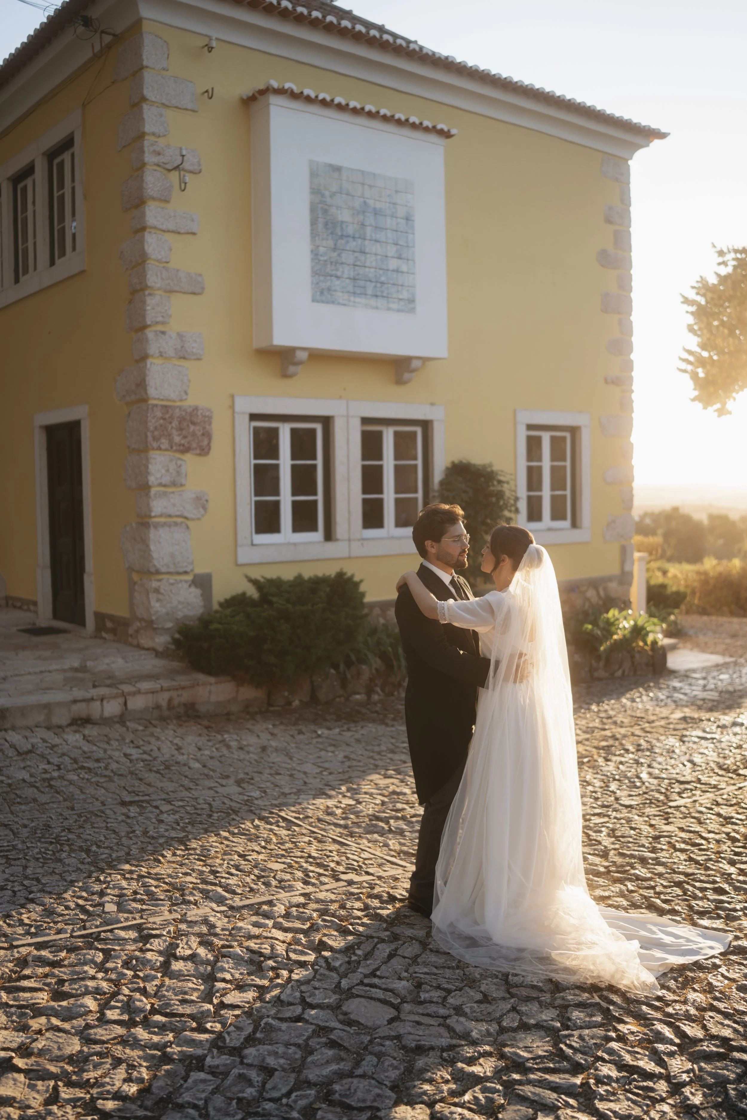 A bride and groom embrace outside on a cobblestone street near a yellow house during sunset.