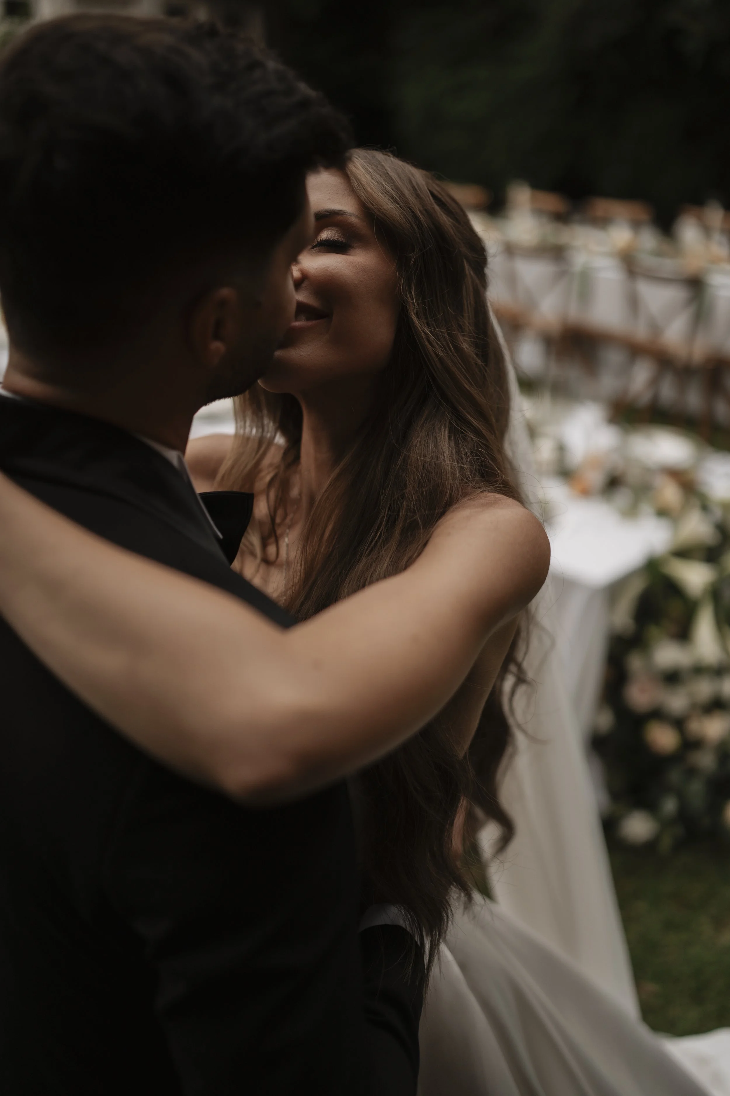A couple sharing a kiss, with a wedding setup in the background, at an outdoor wedding reception.