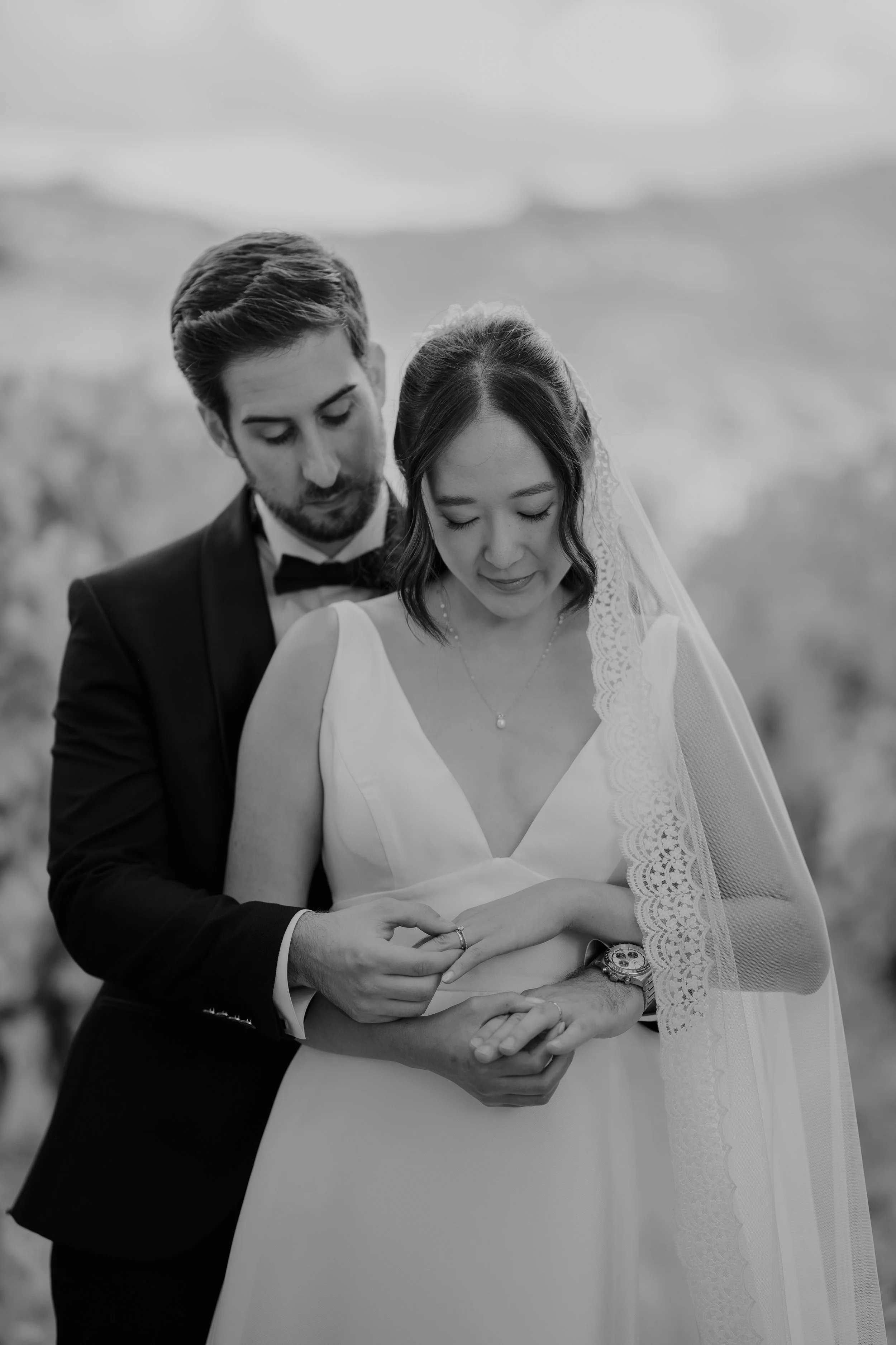 A black-and-white photo of a bride and groom holding hands, with the groom placing a ring on the bride's finger. The groom is dressed in a tuxedo with a bow tie, and the bride is wearing a white wedding gown with a lace veil and a necklace.