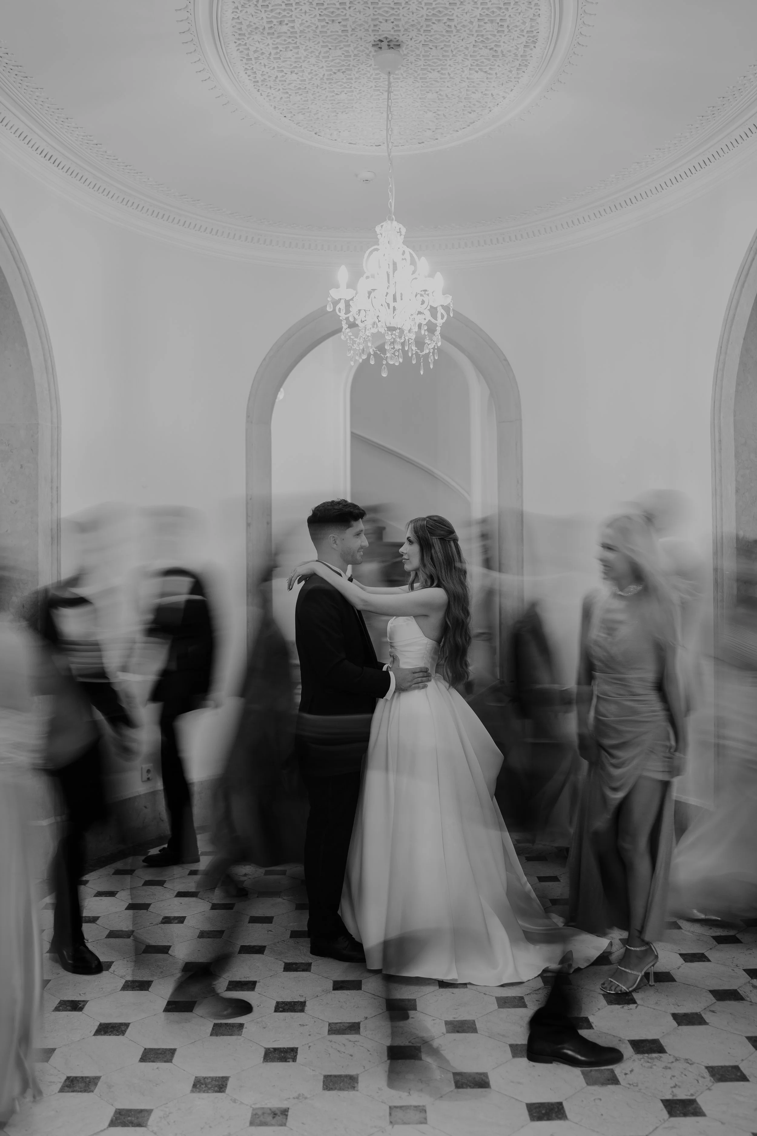 A bride and groom dancing in the center of a ballroom, surrounded by blurred guests, under a chandelier and ornate ceiling.