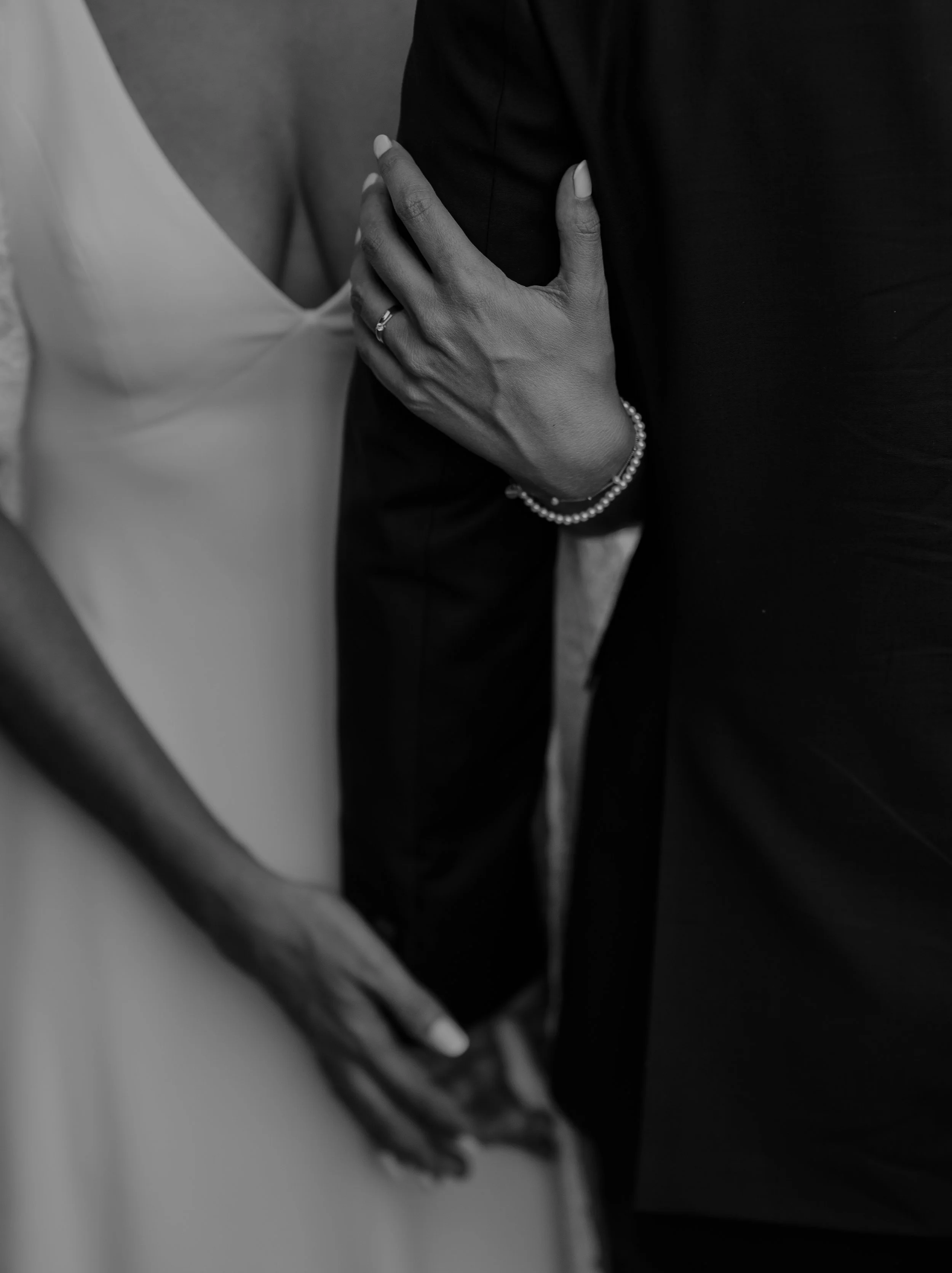 Close-up of a bride and groom holding hands during a wedding ceremony, with the bride wearing a wedding dress and the groom in a dark suit, in black and white.