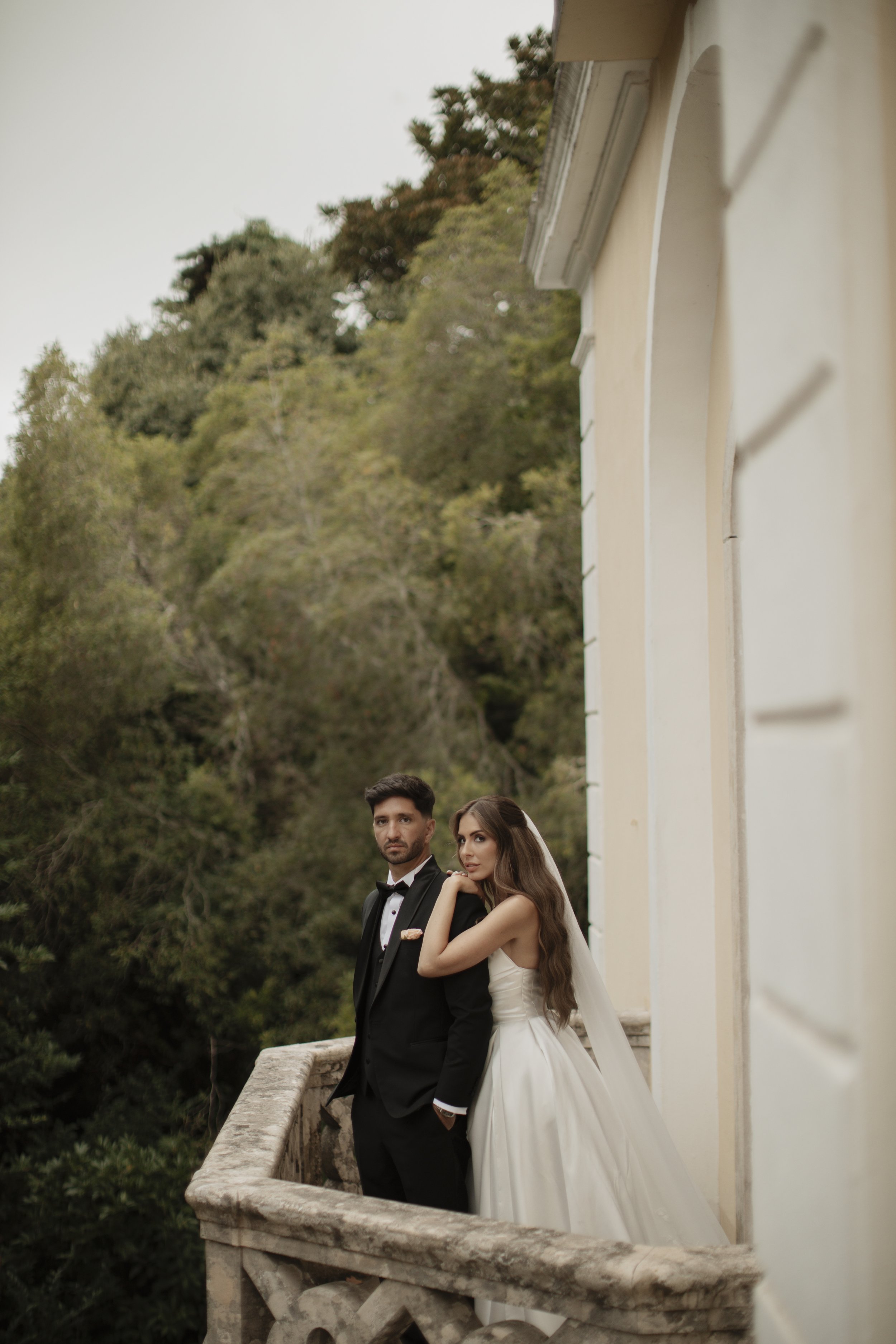 A bride and groom standing on a balcony outdoors during daytime with green trees and a hillside in the background. The groom is wearing a black tuxedo with a bowtie, and the bride is in a white wedding gown with a veil. The bride is resting her arm o