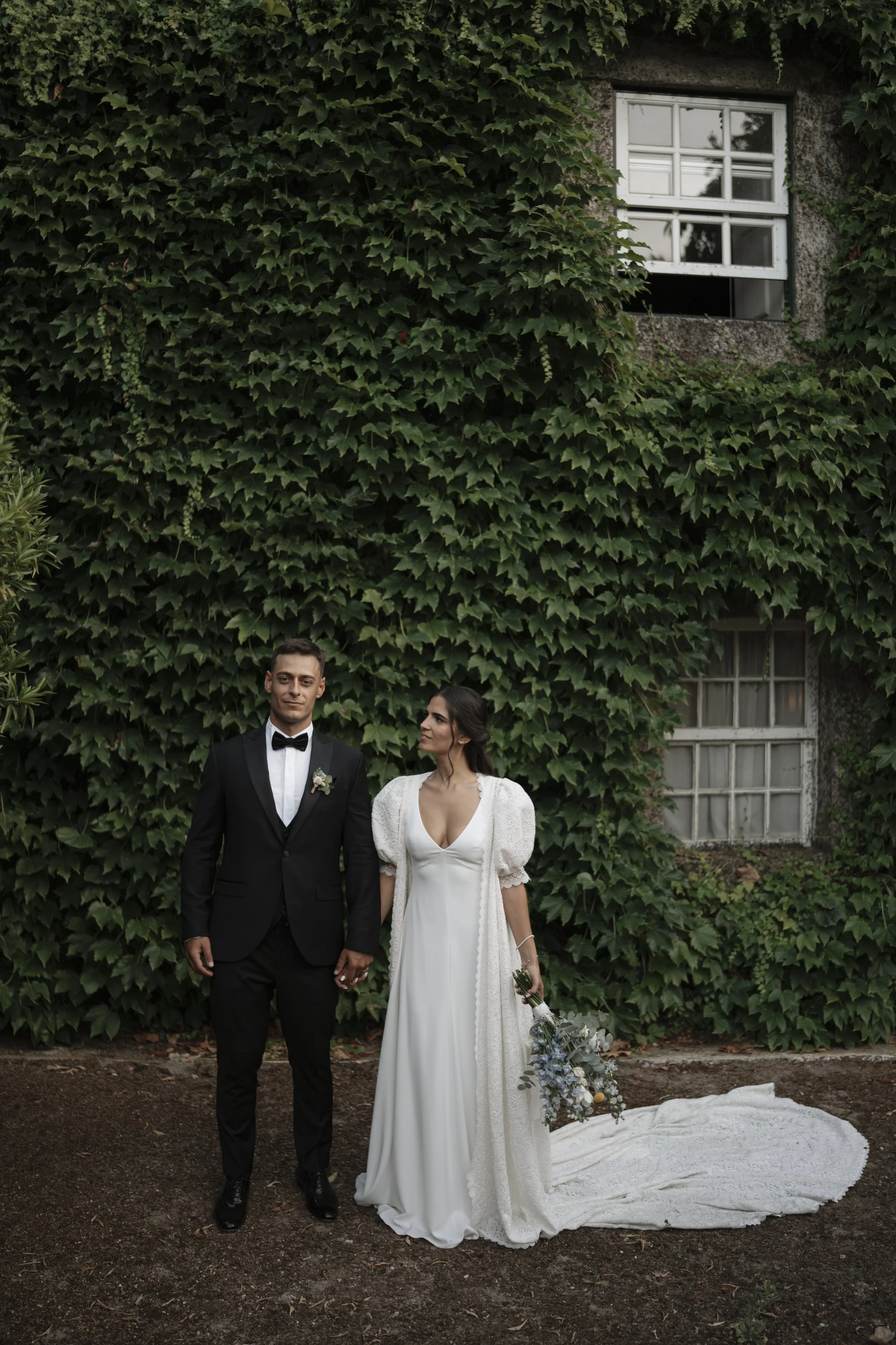 A bride and groom holding hands in front of a ivy-covered wall, with an open window in the background. The groom is in a black tuxedo with a bow tie, and the bride is in a white wedding dress holding a bouquet.