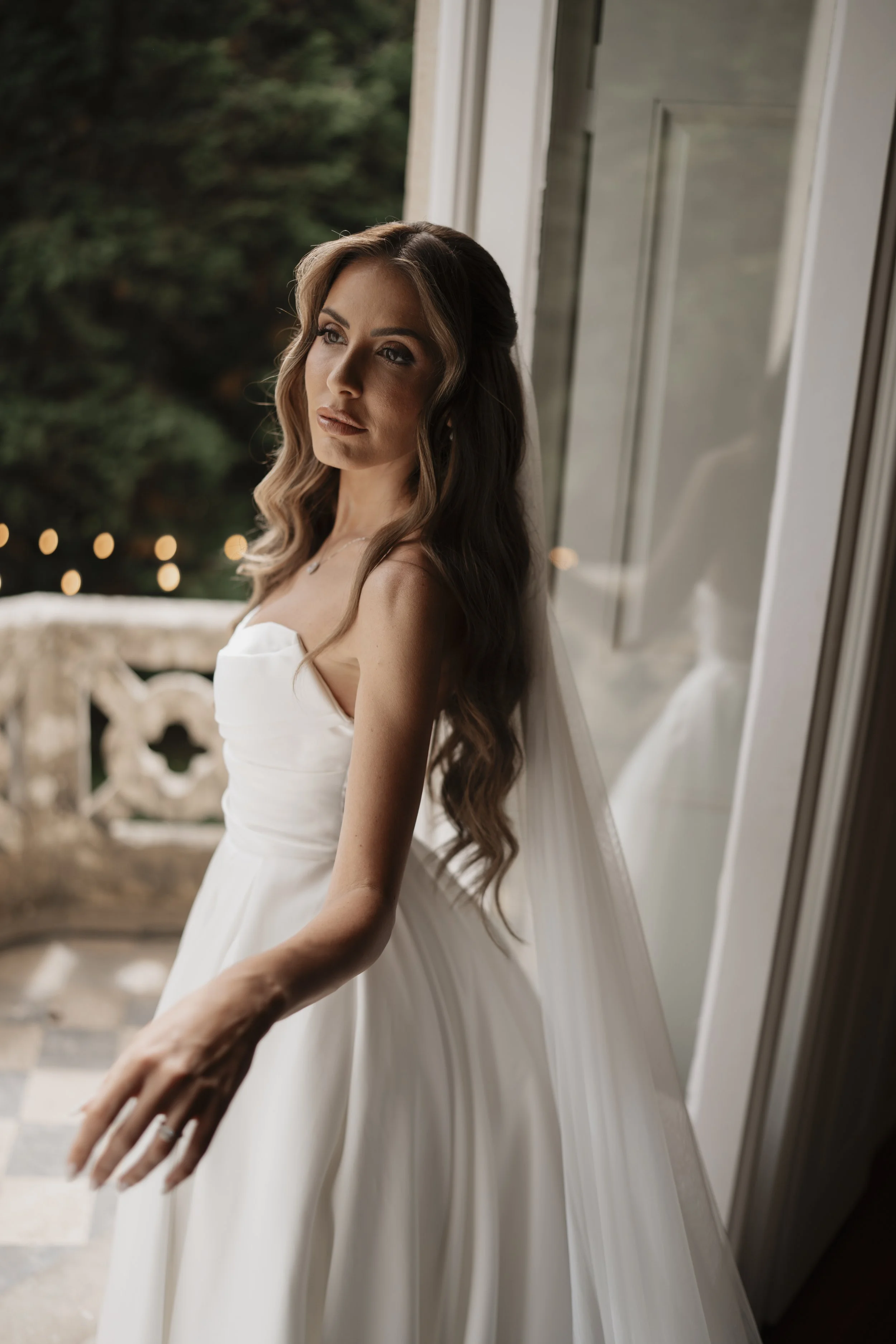A bride in a white wedding dress standing by a window, gazing outside with a contemplative expression.