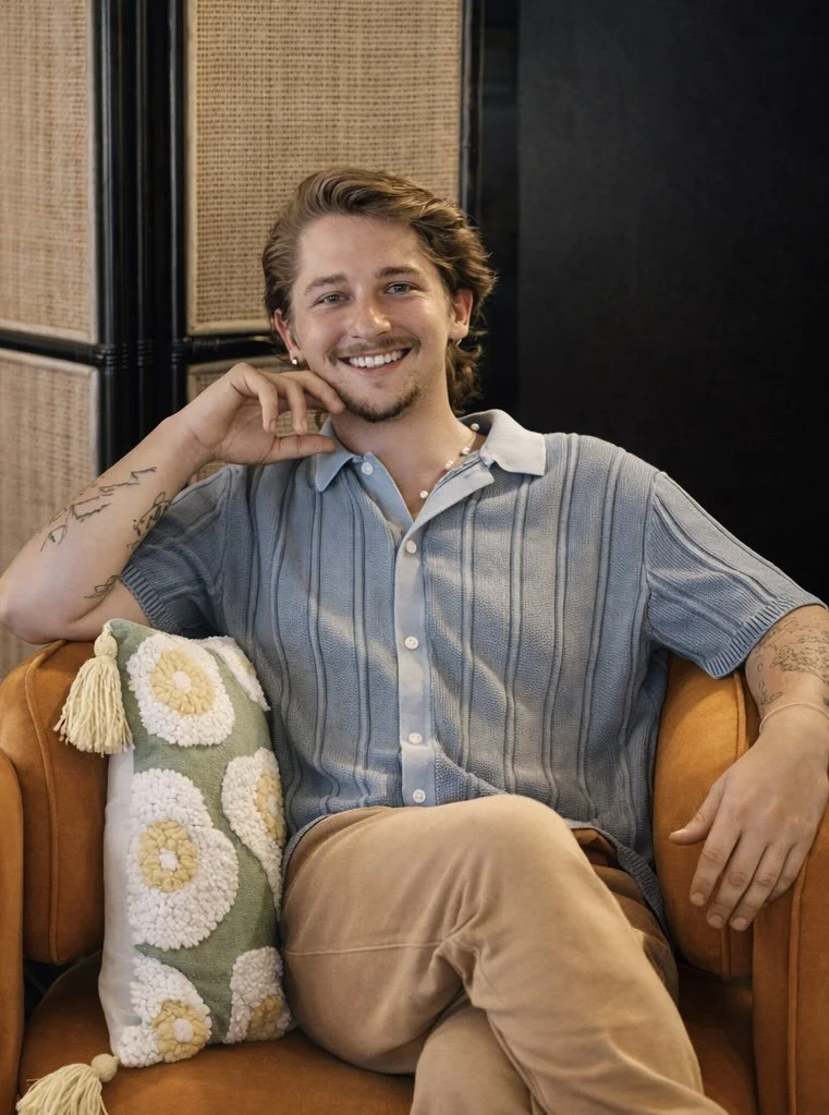 Smiling young man with wavy hair and tattoos sitting on an orange sofa next to a decorative pillow.