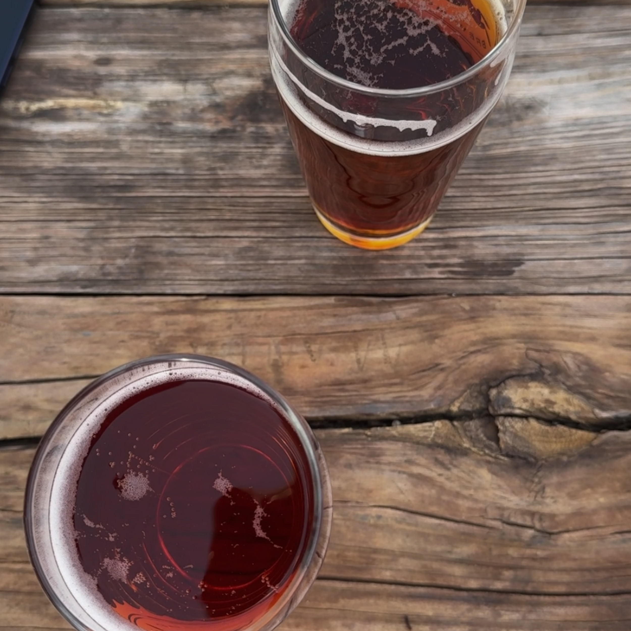 A close-up photo of a glass of dark red wine on a wooden table, with the wine's surface showing bubbles and slight foam. The image is taken from above, showcasing the drink inside the glass.