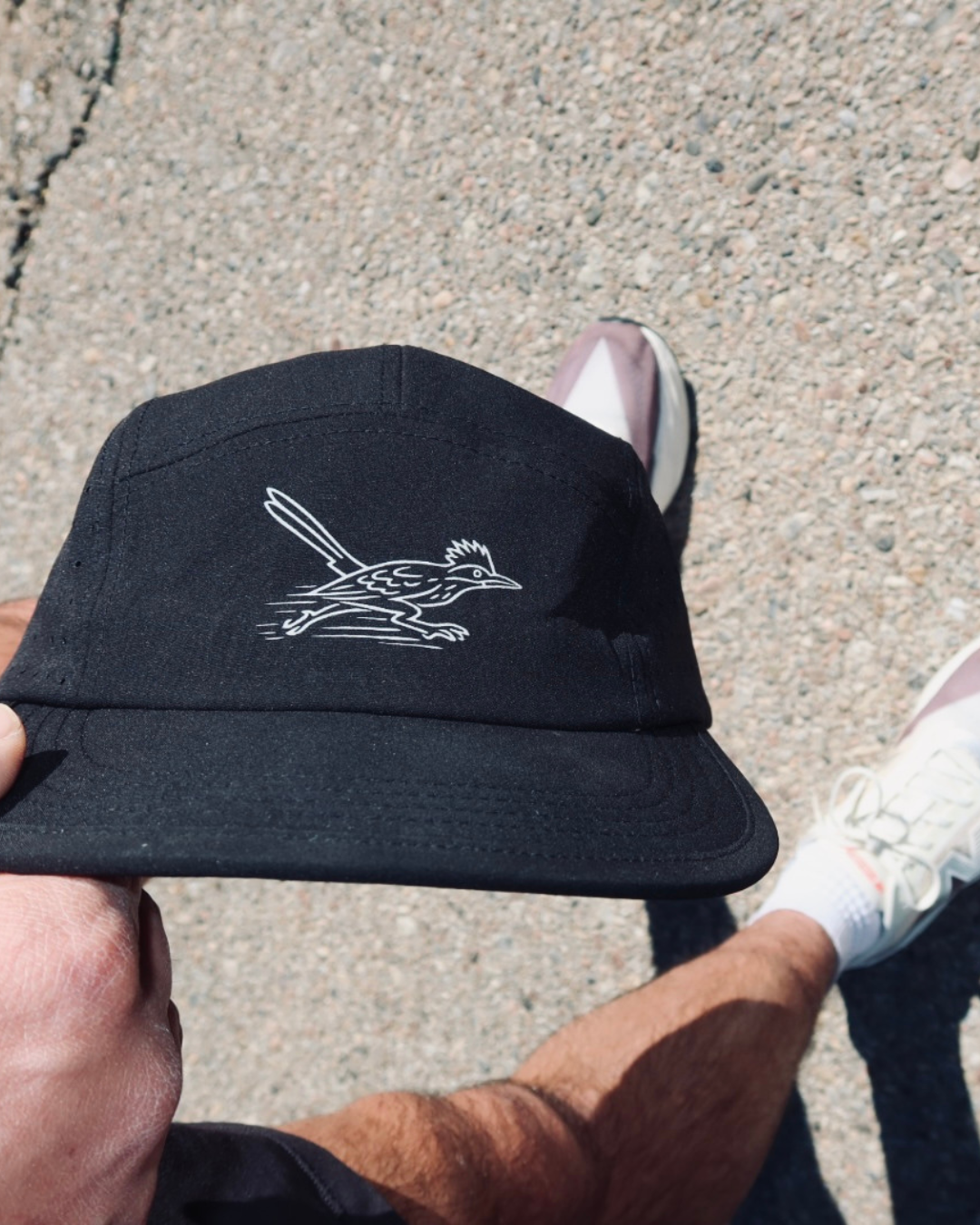Person holding a black cap with a white bird logo, standing on a gravel surface, wearing white sneakers and black shorts.