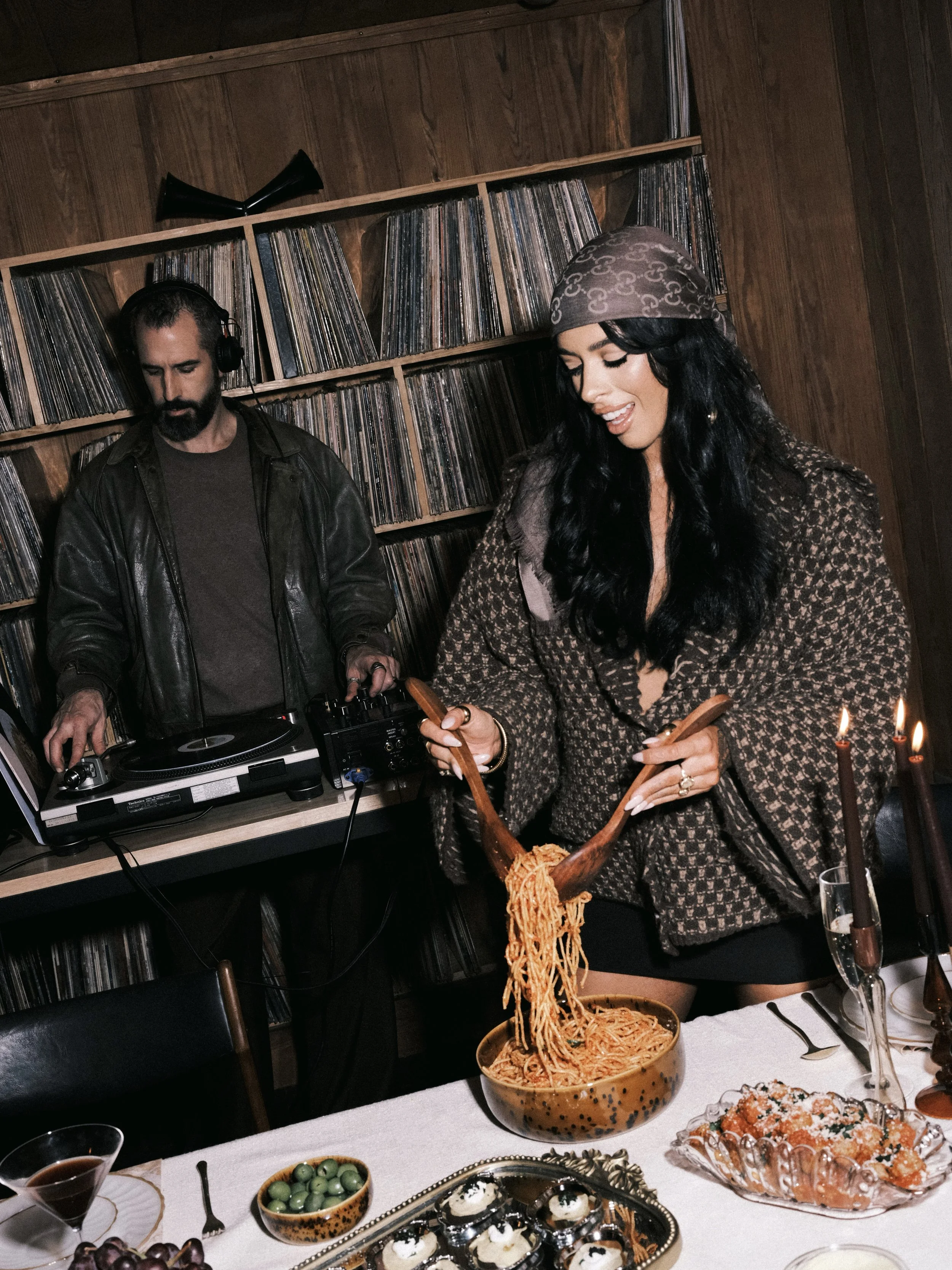 A woman with long dark hair pulling spaghetti from a bowl at a dinner table, with a DJ playing music in the background.