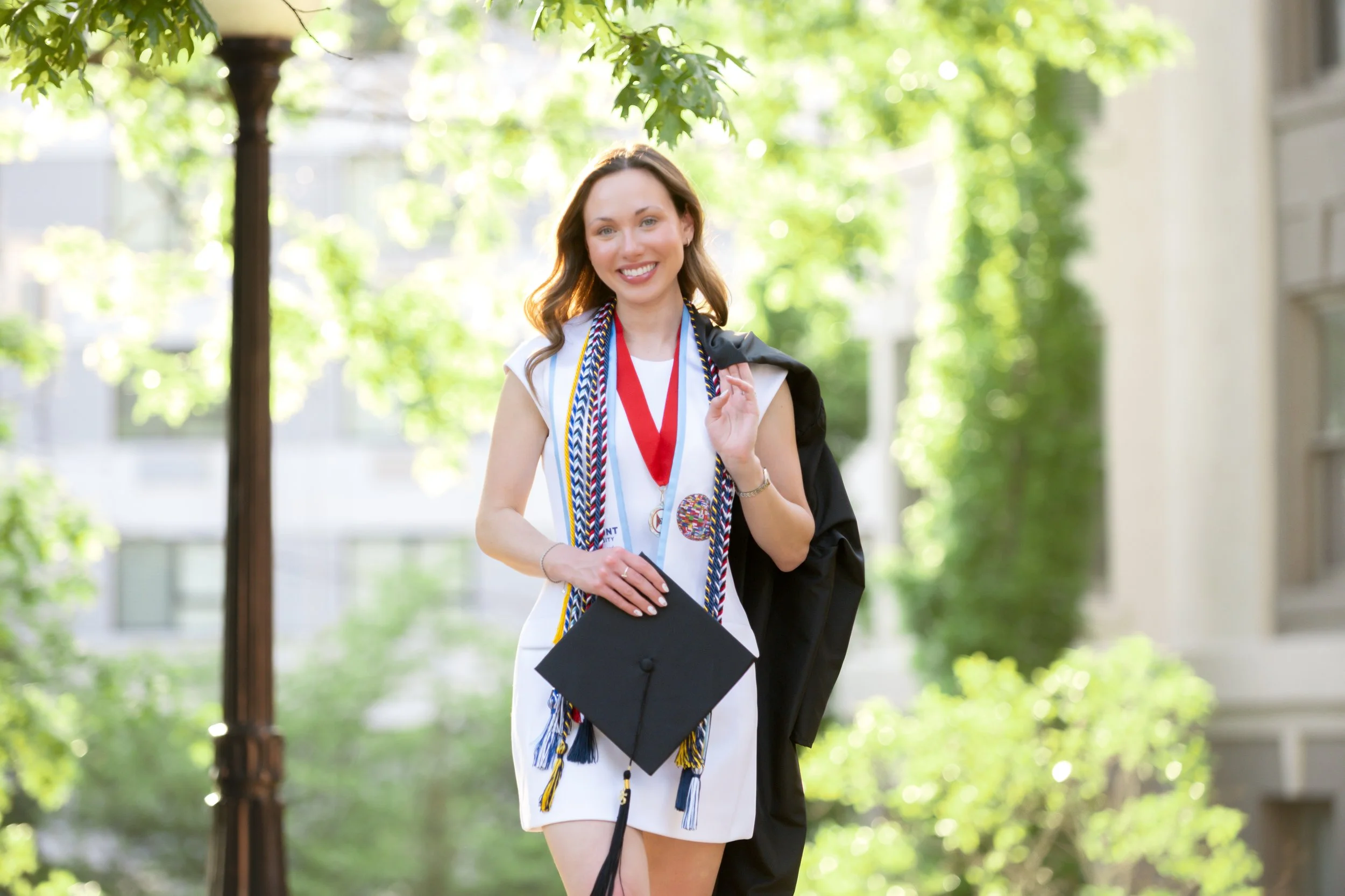 Young woman in a white dress smiling and holding a graduation cap, with several honor cords and medals around her neck, outdoors in a sunny park area.