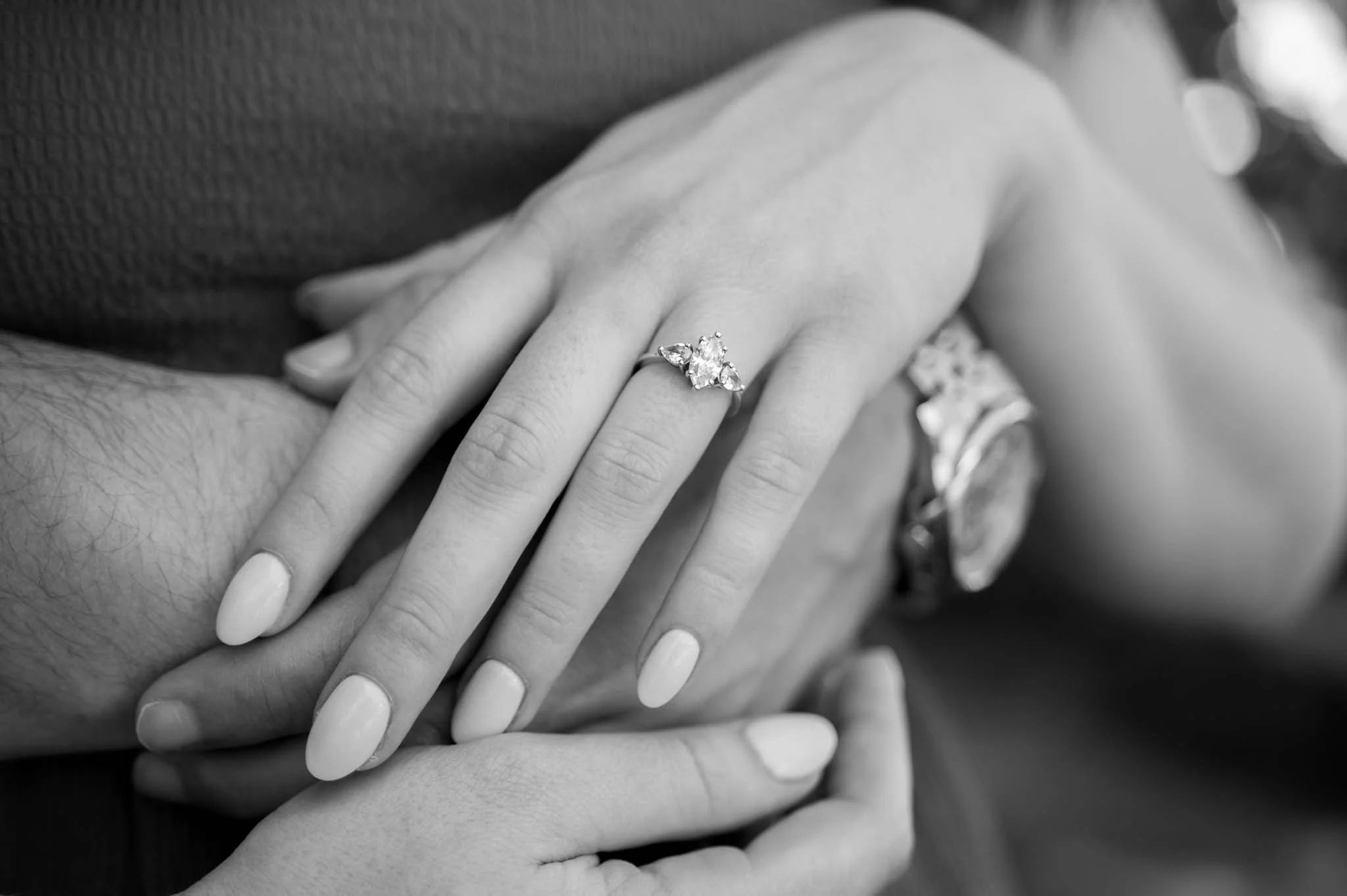 Close-up of a woman's hand with an engagement ring featuring a large central gemstone and smaller side gemstones, resting on a man's hand, both hands with neatly manicured nails. The woman's other wrist has a watch. The background is blurred.