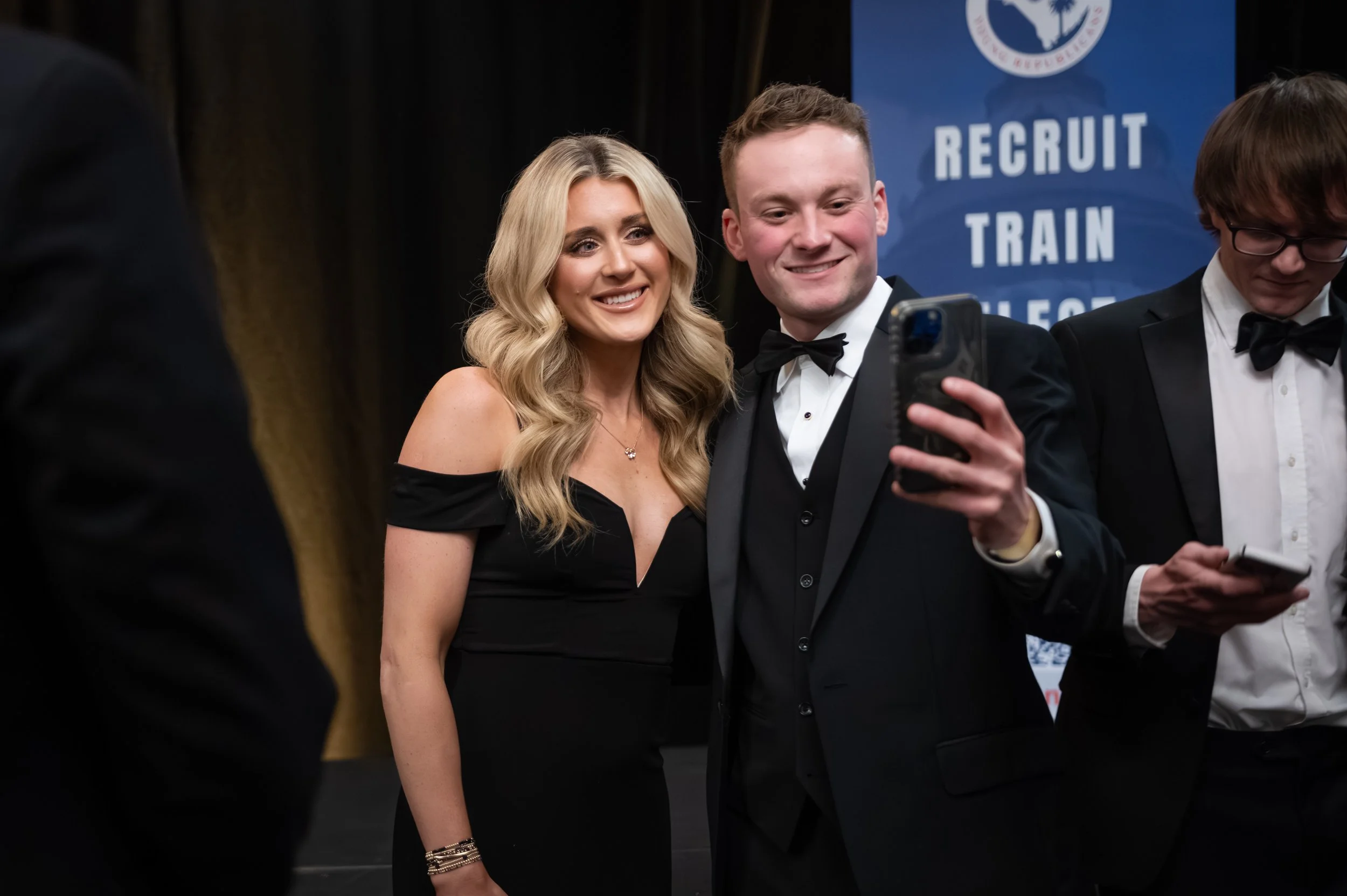 Two people in formal attire taking a selfie at an event with a sign that reads "Recruit Train" in the background.