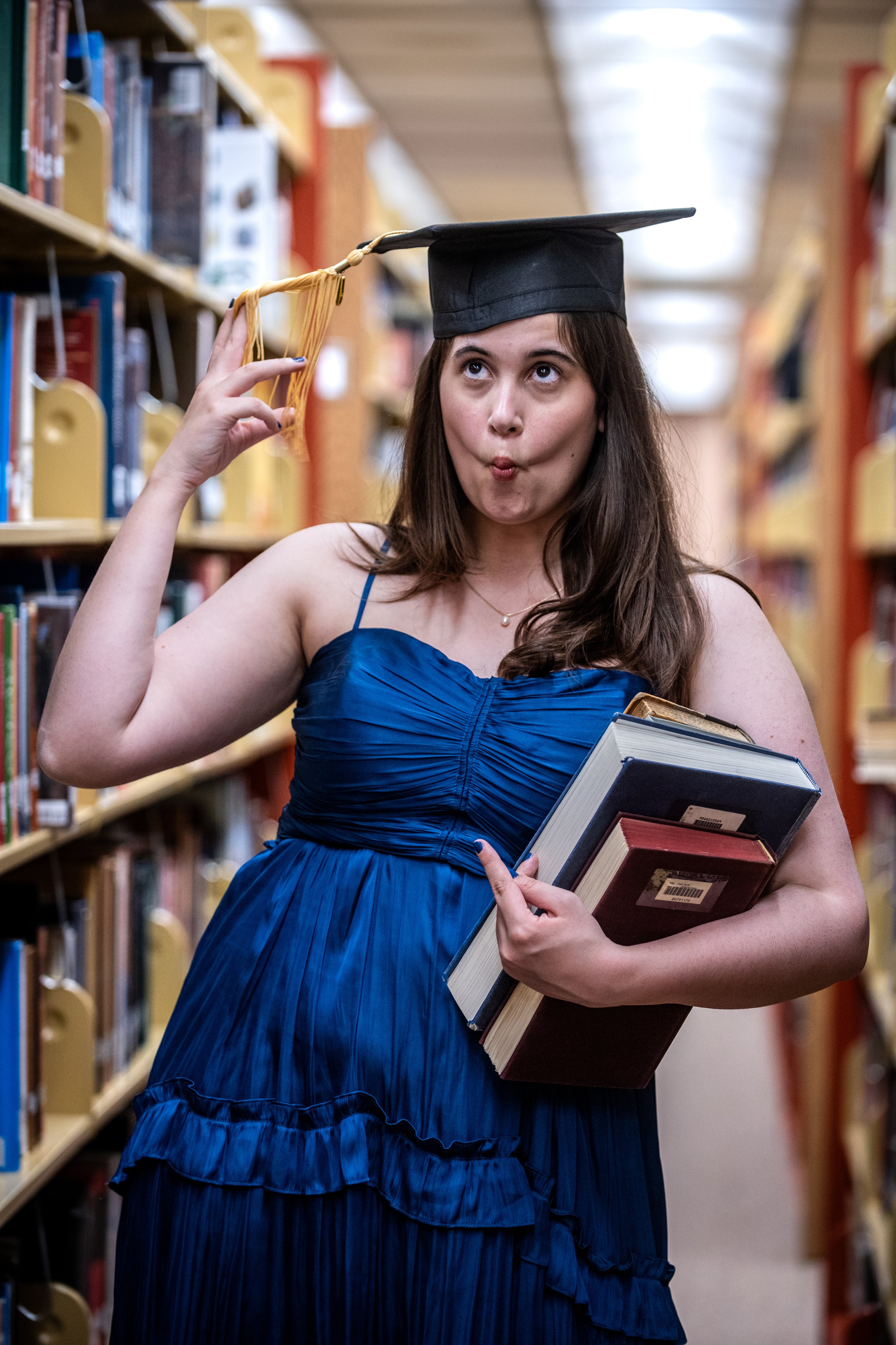 A young woman in a blue dress and graduation cap holding books in a library, making a playful face and posing with her hand near her head.