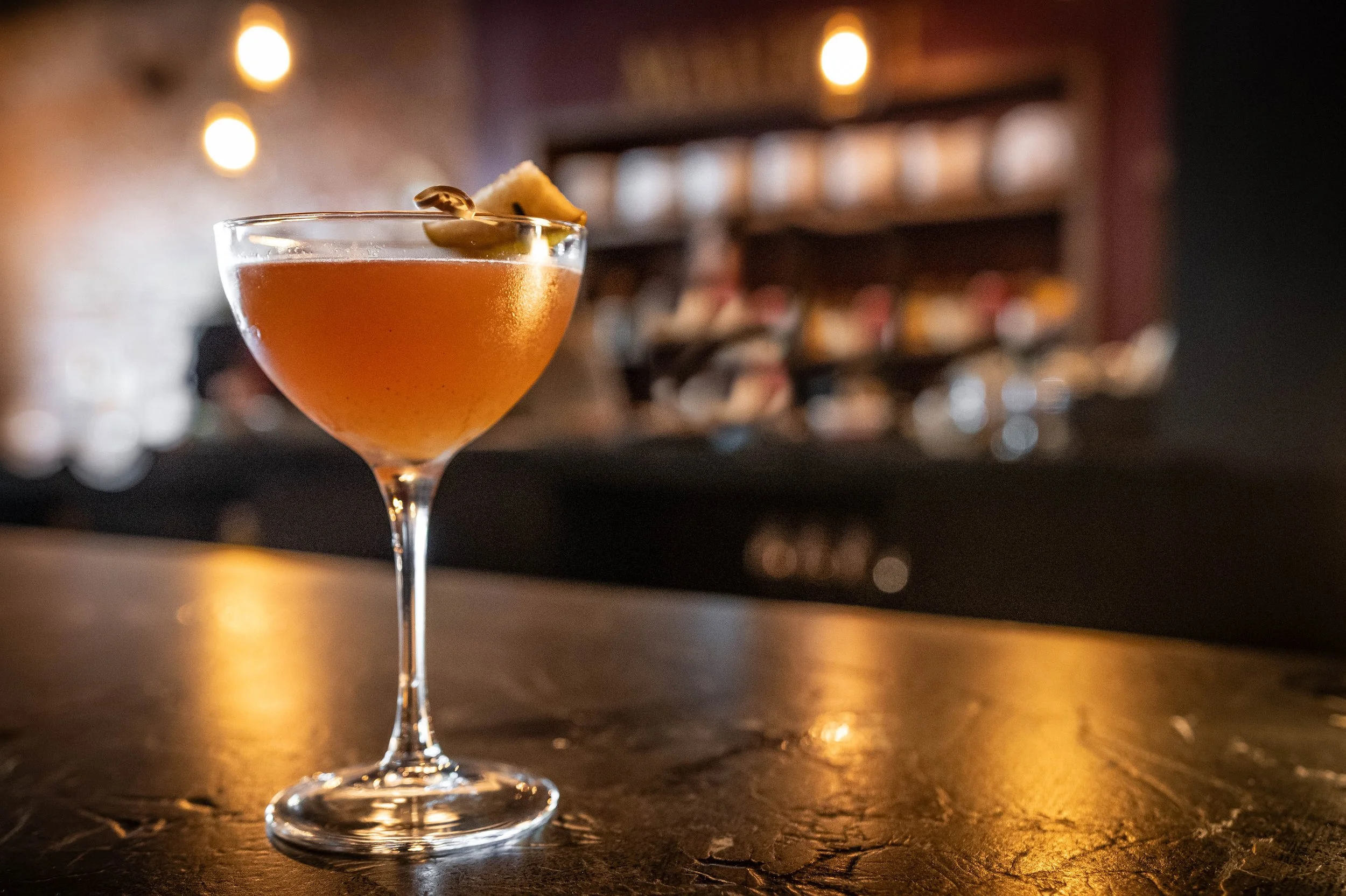 A cocktail in a coupe glass with an orange or amber-colored drink, garnished with a nut or small piece of garnish, set on a wooden bar counter in a dimly lit bar or lounge.
