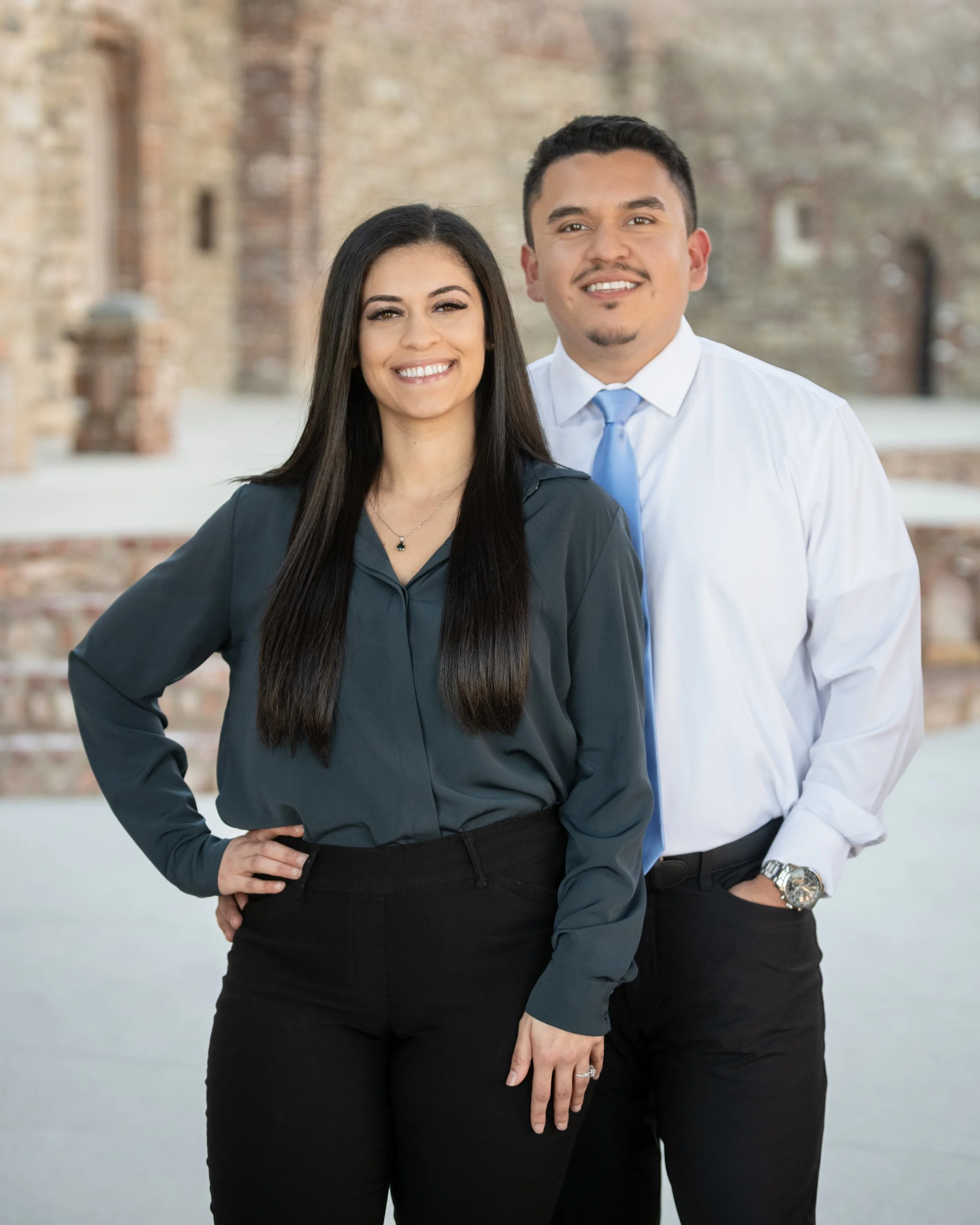 A smiling woman with long dark hair and a man in a white shirt with a tie standing outdoors with an old brick building in the background.
