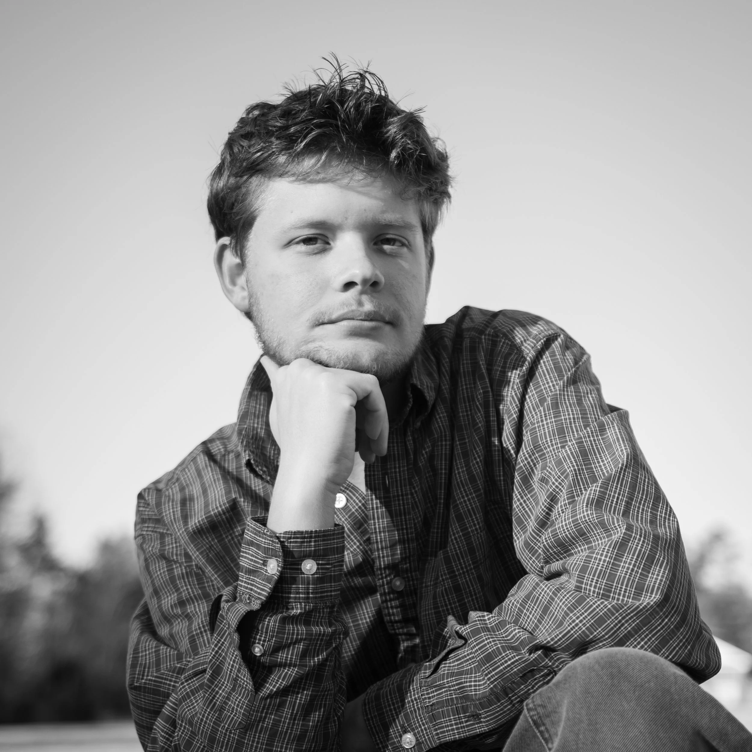 Black and white photo of a young man with short, tousled hair, wearing a checkered shirt, resting his chin on his hand and looking at the camera with a serious expression.