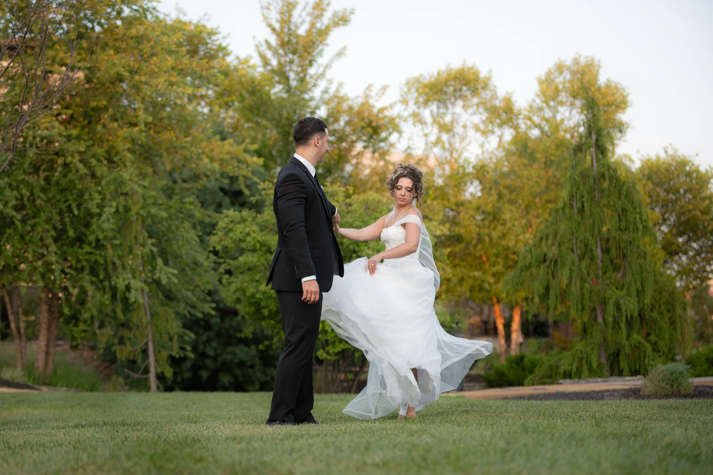 A bride and groom dance outdoors on a grassy area with trees in the background during sunset.