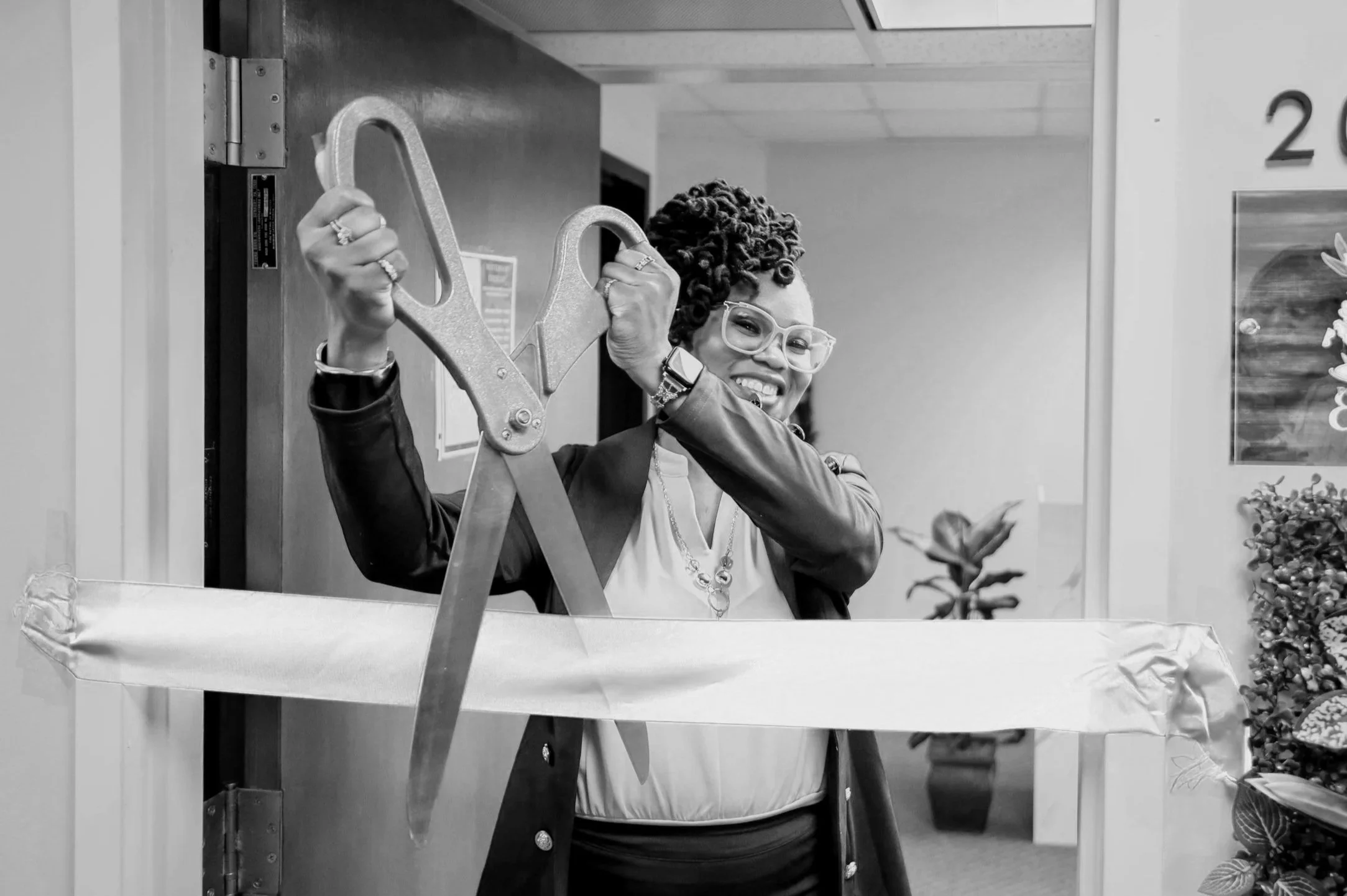 A woman with curly hair and glasses is smiling and holding a large pair of scissors over a ribbon at a ribbon-cutting event.